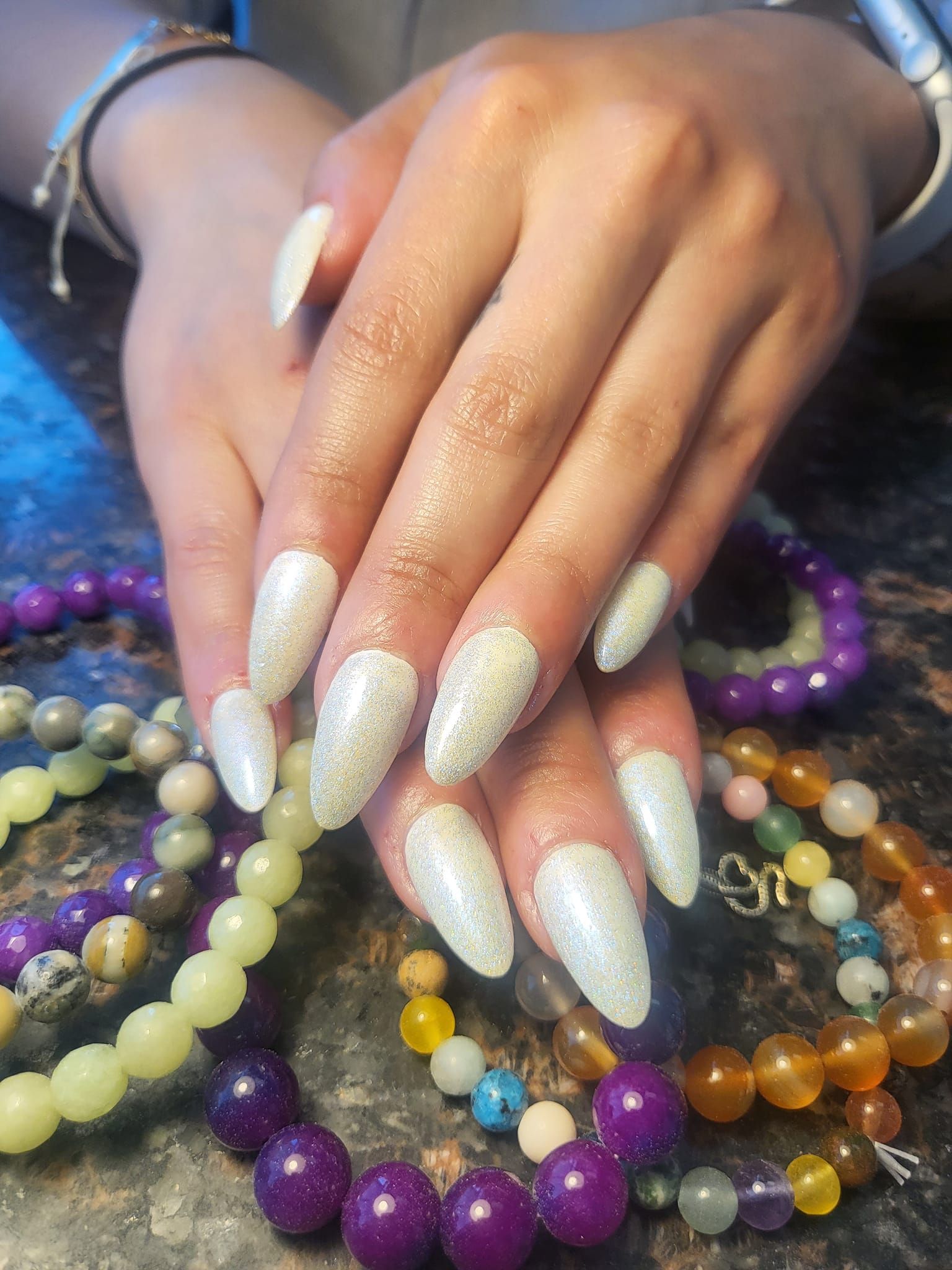 Woman's hands with long, almond-shaped white glitter nails, resting above colorful beaded bracelets on a table.