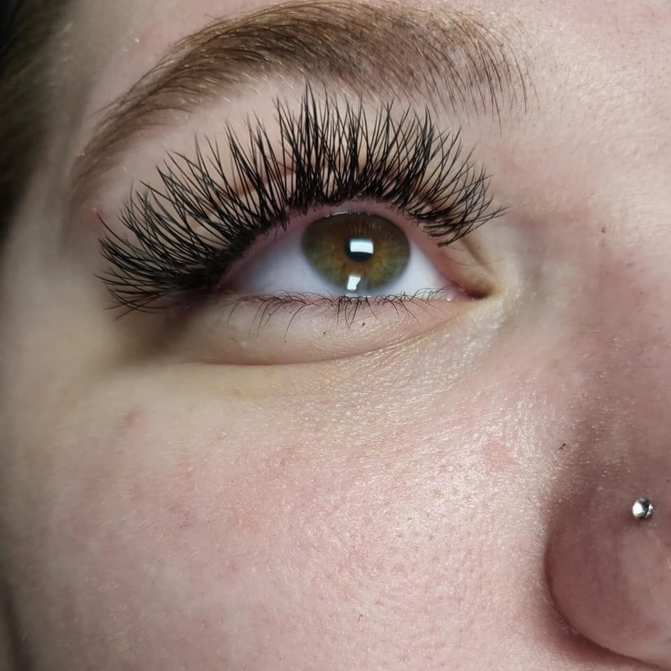 Close-up of an eye with lush, dark lash extensions. The eye is hazel and the skin is fair, with a nose piercing.