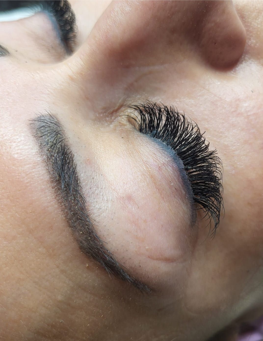 Close-up of a person's closed eye with dark, thick eyelash extensions and a dark, filled-in eyebrow.