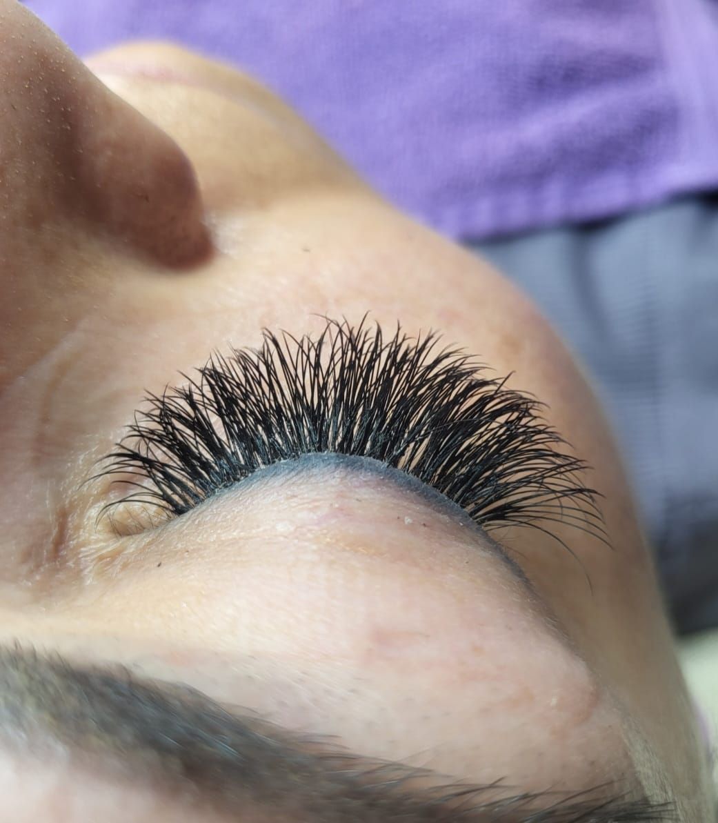 Close-up of a person's closed eye, showcasing dense, dark eyelash extensions. A light-colored eyebrow is also visible.