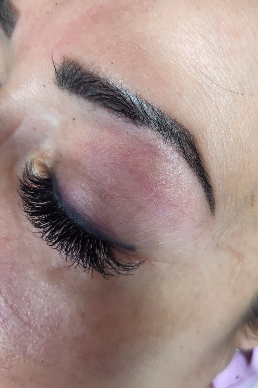Close-up of a person's eye with thick, dark lashes, a filled-in eyebrow, and purple eyeshadow.