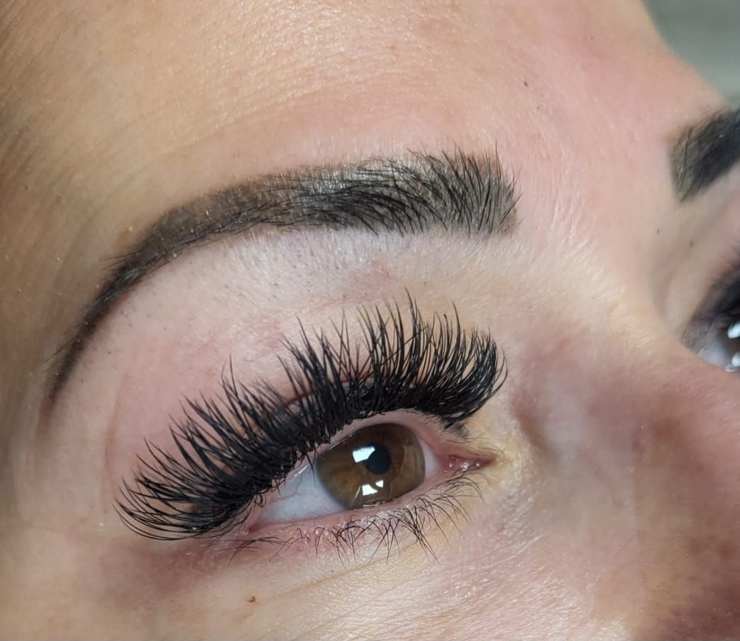 Close-up of an eye with dark, full eyelash extensions and sculpted eyebrows. The eye is brown.