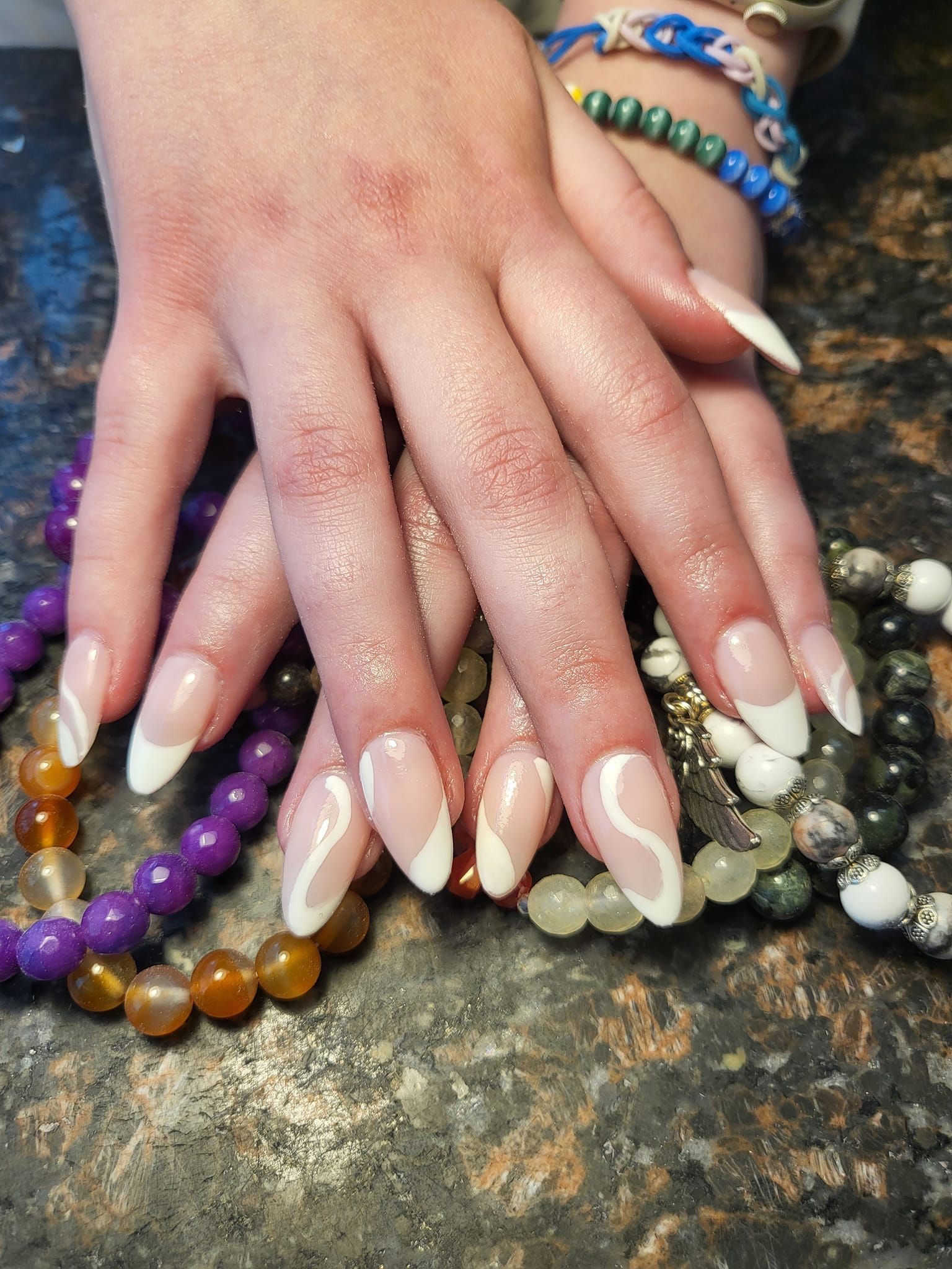 Hands with long, almond-shaped nails. Clear polish with white french tips and wavy white designs, resting on a beaded surface.
