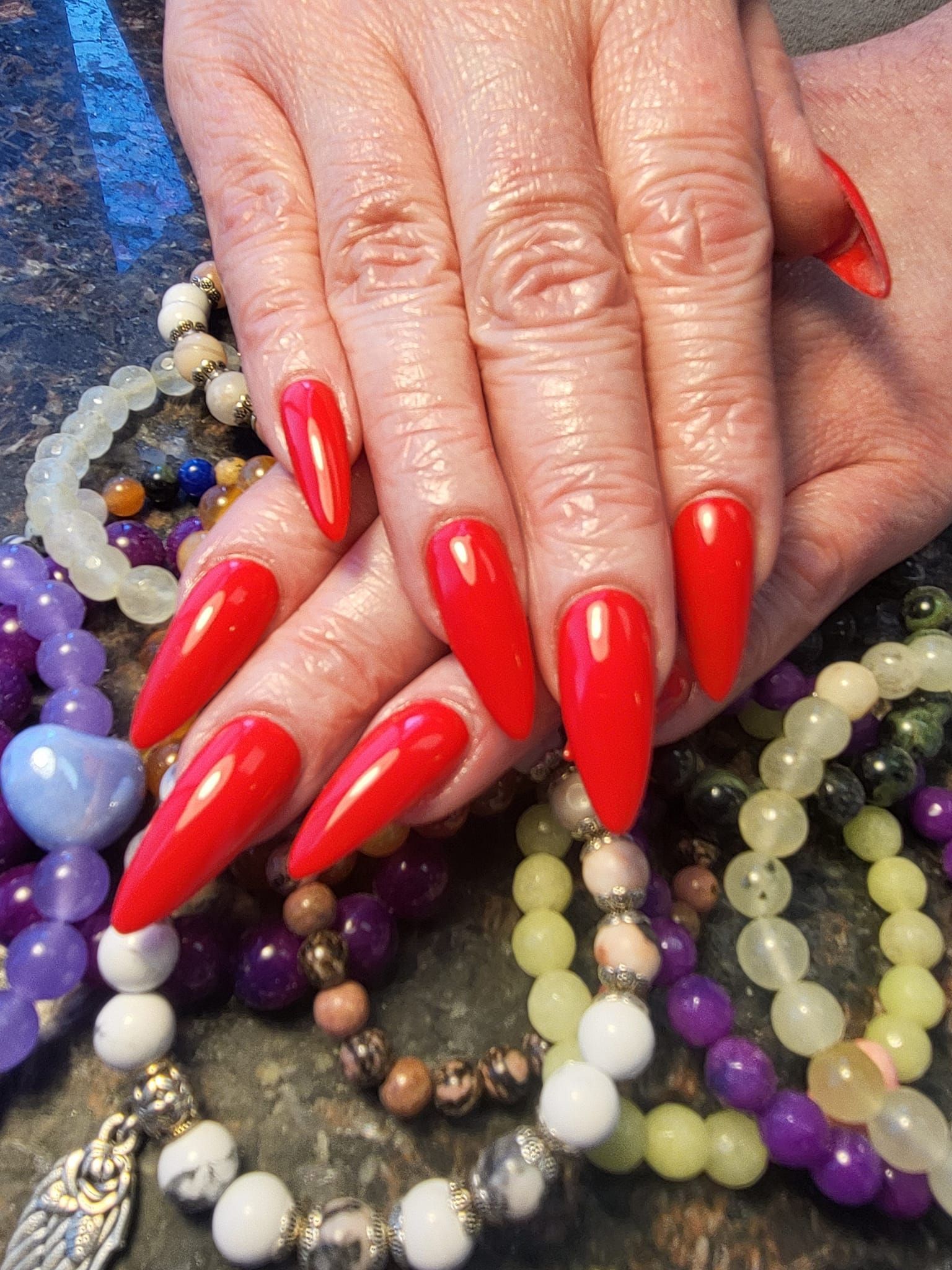 Hand with long, pointed red fingernails, resting on a bed of colorful beaded jewelry.