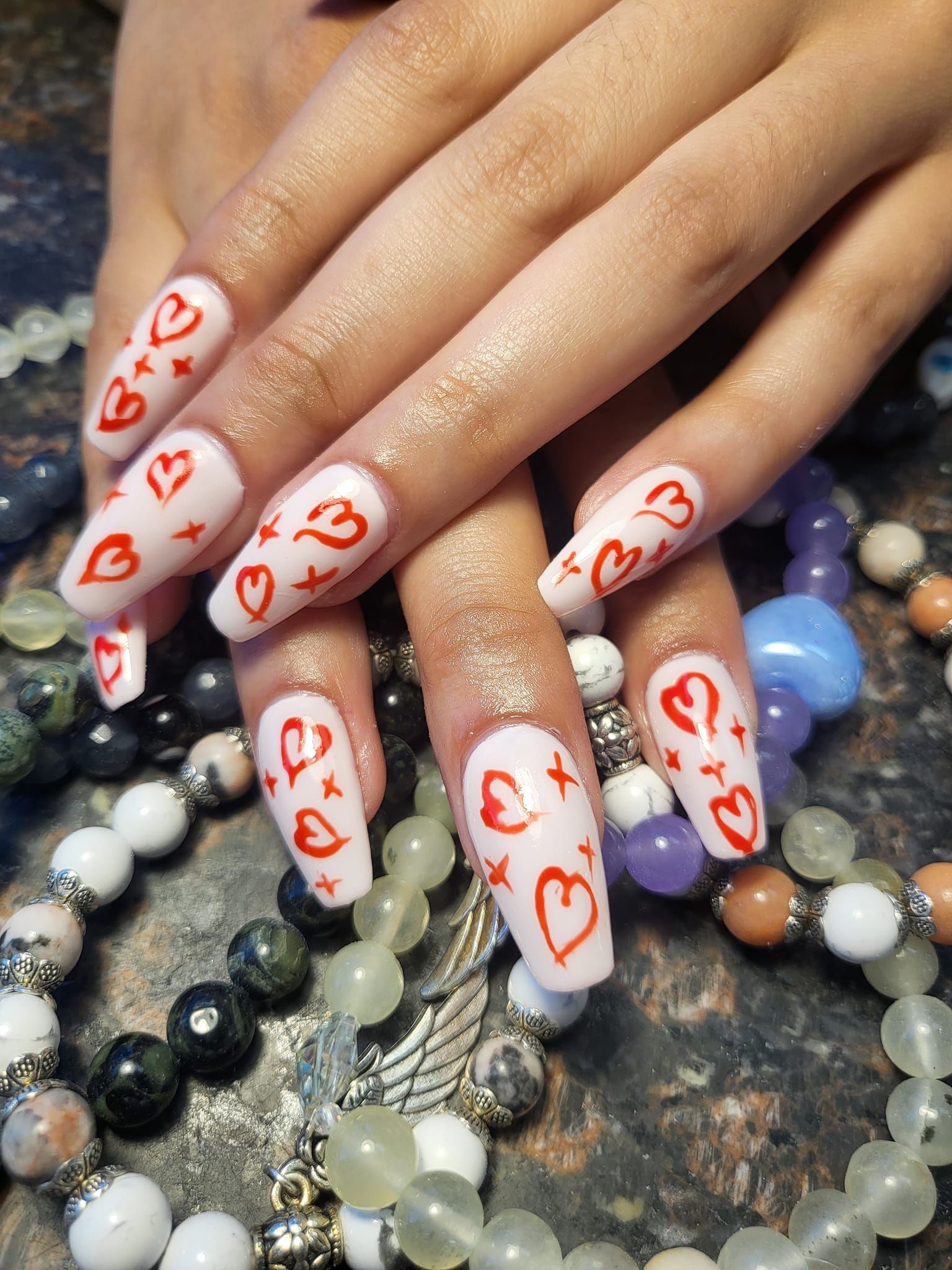 Hands with white coffin nails decorated with red hearts and stars, resting on beaded jewelry.