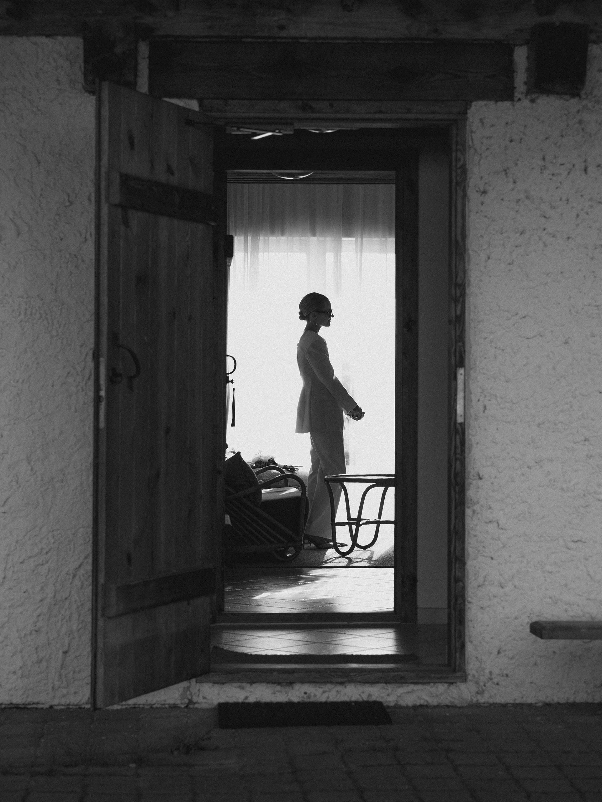 Person silhouetted in a doorway; inside, a sunny room with sheer curtains; exterior view.