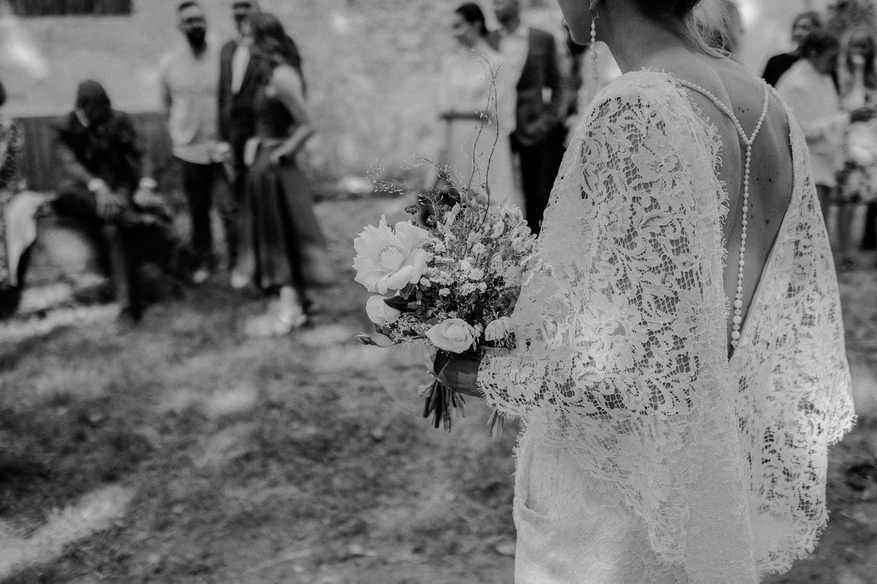Bride in lace dress holding flowers, back to camera, outdoors with gathered guests.