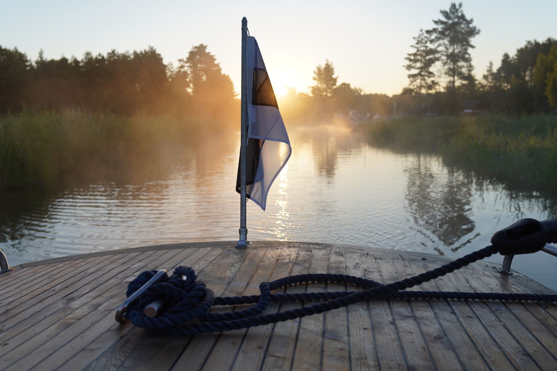 Boat on a misty river at sunrise, with a flag and a rope on deck.