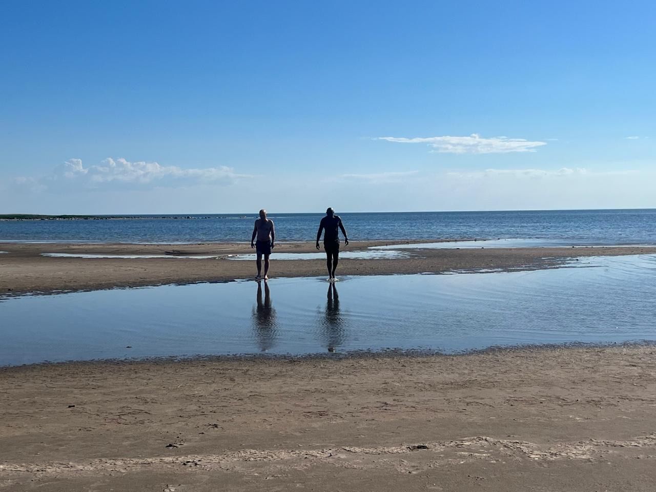 Two people walking in shallow water towards the ocean on a sunny beach. Blue sky.