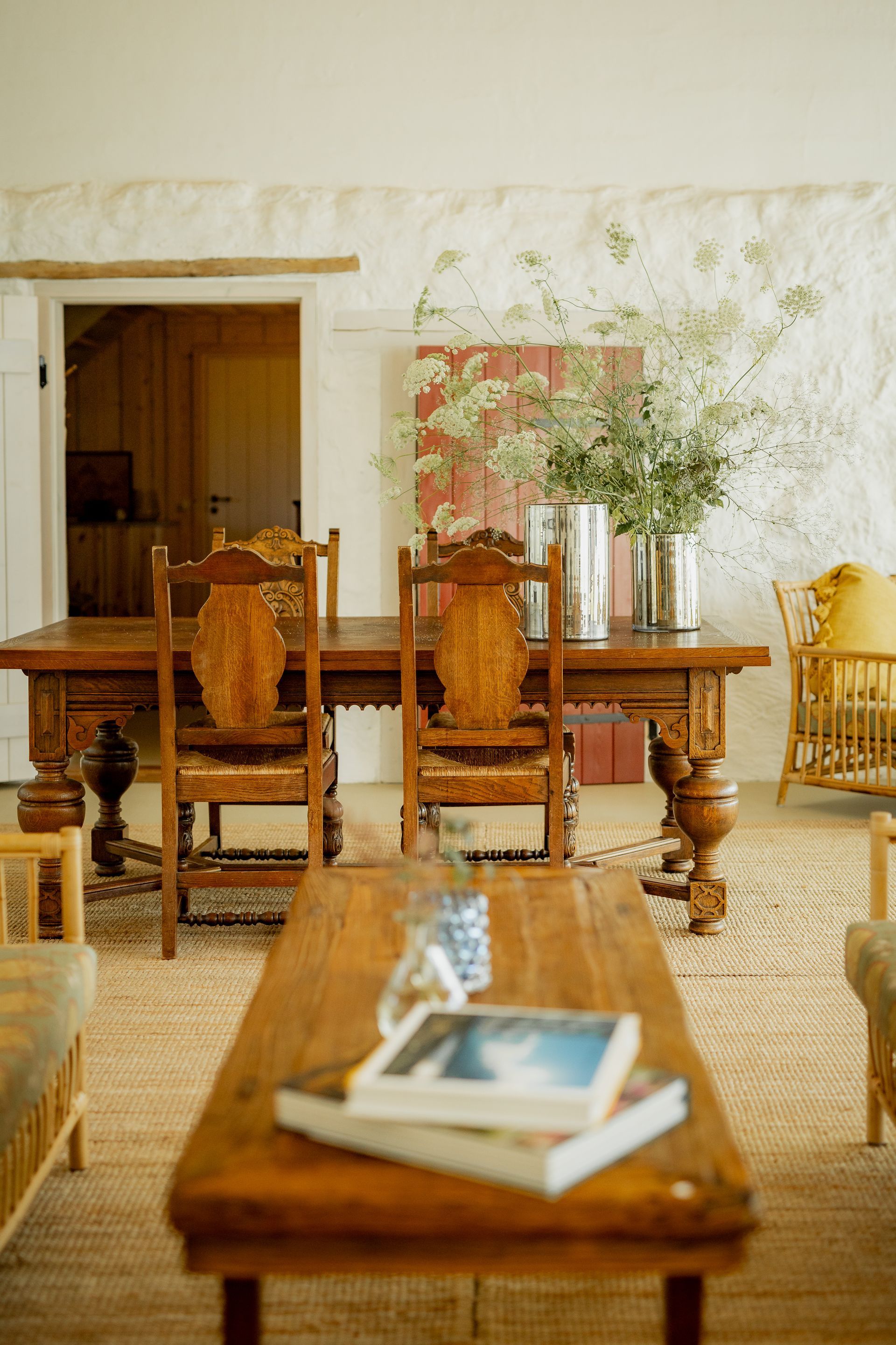 Rustic wooden dining table and chairs in light-filled room, flowers in vase, and a coffee table with books.