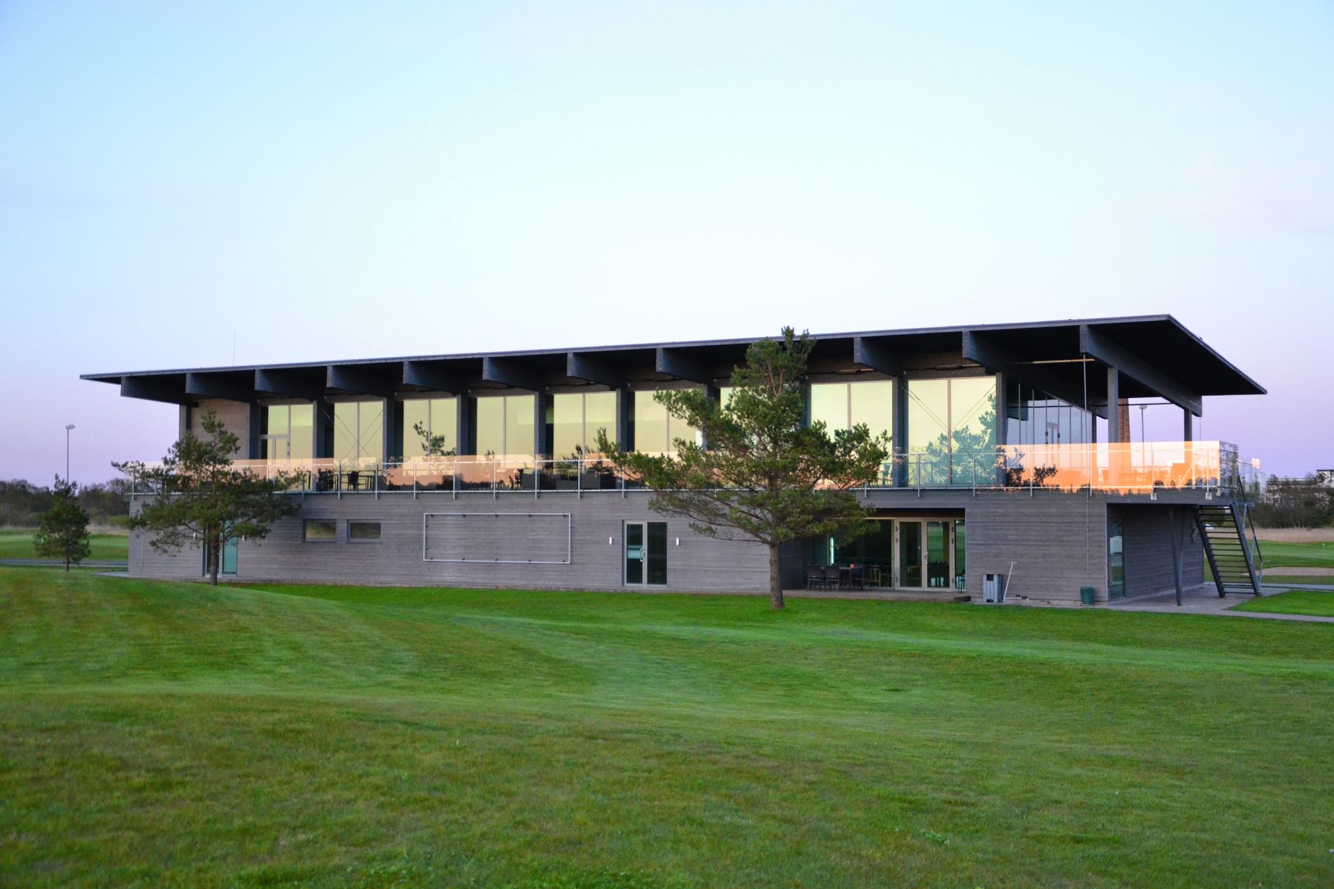 Modern two-story building with large windows and a flat roof, set on a green grassy hill, possibly a golf course clubhouse.