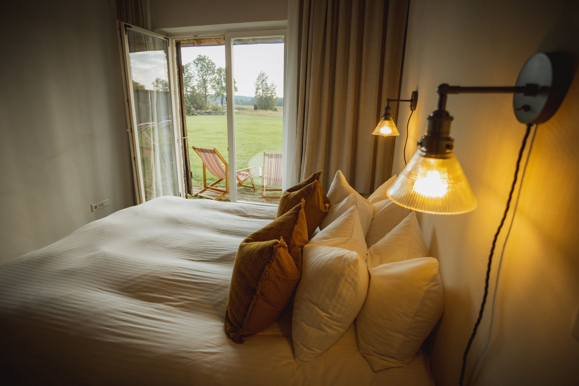 Bedroom with unmade bed, pillows, wall lamps, and open door to a grassy outdoors.