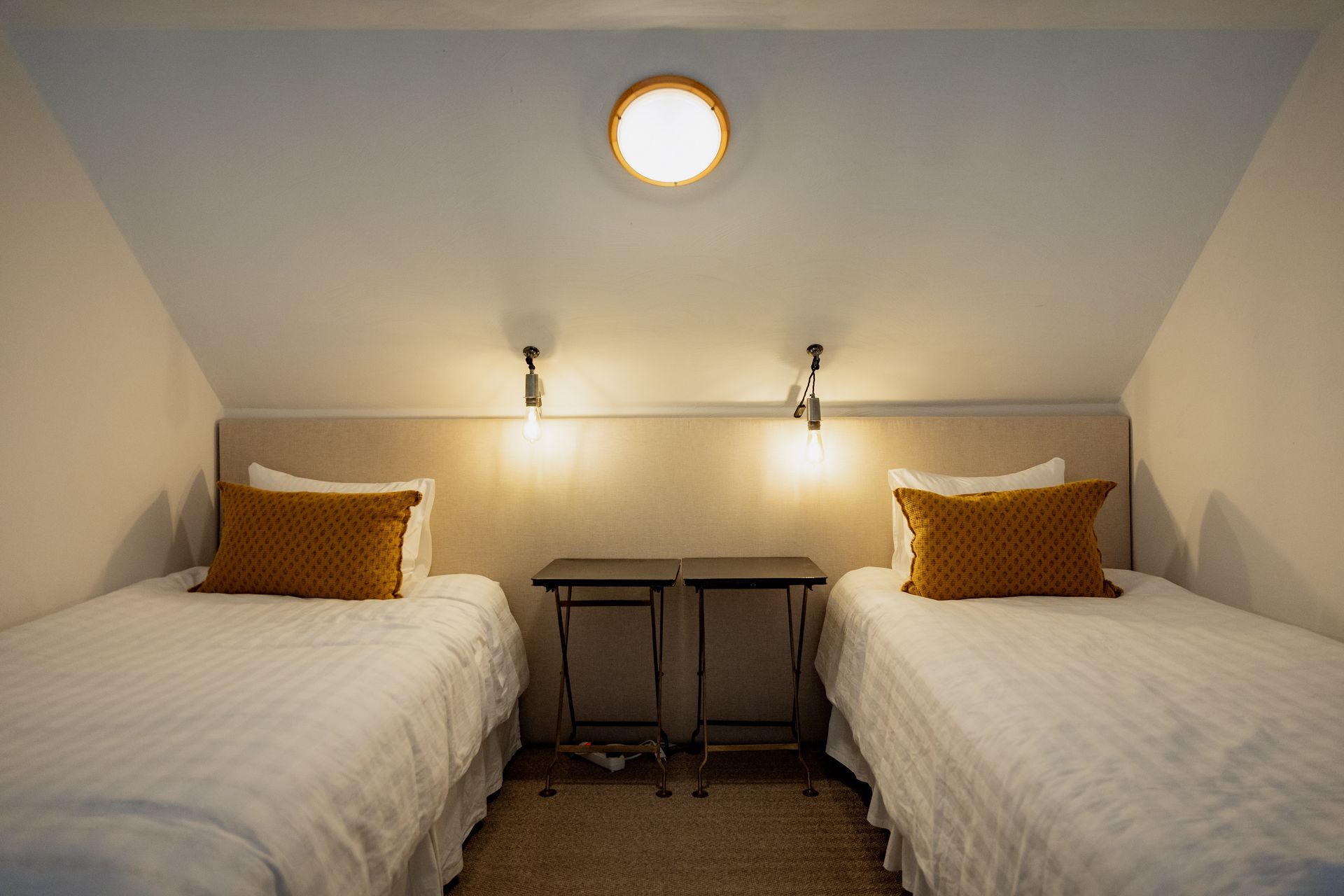 Two twin beds with mustard pillows, lit by wall sconces and a central ceiling light in a neutral-toned room.
