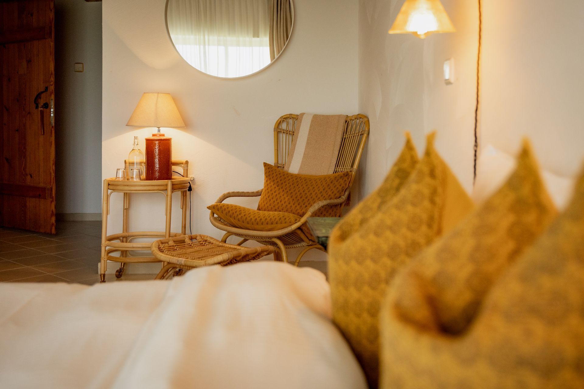 Bedroom with woven chair, side table with lamp, large bed with yellow pillows, and a round mirror.