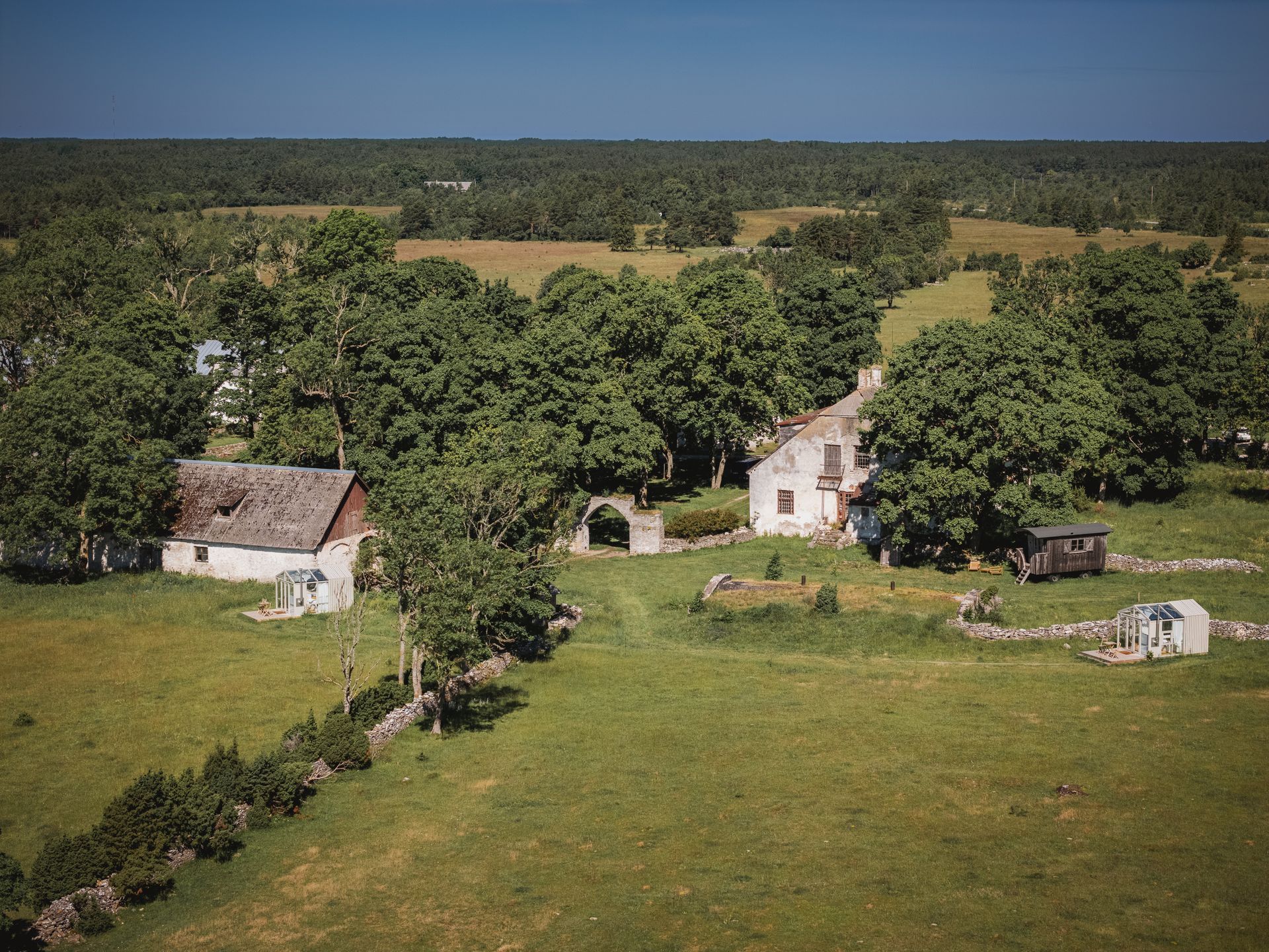 Lush green field with stone fences, trees, and several white buildings with brown roofs under a clear blue sky.