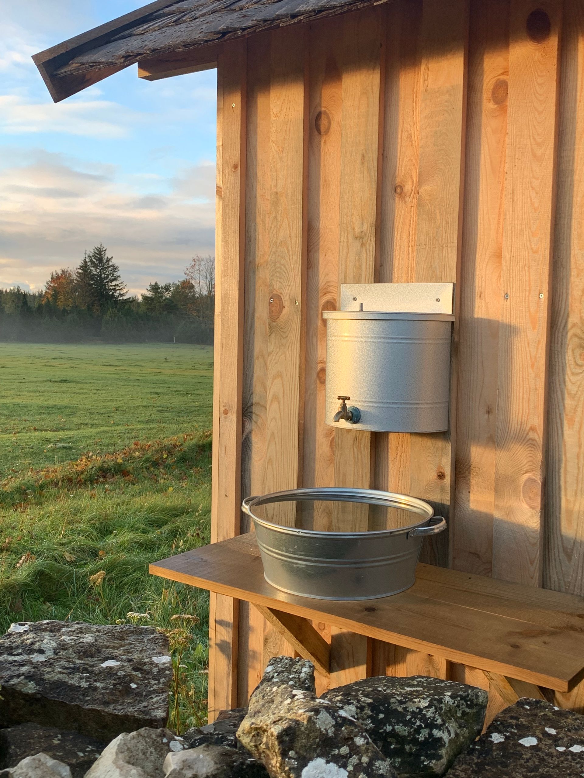 Galvanized metal washbasin setup outdoors: water dispenser above a basin, set against a wooden wall, with a grassy field in the background.
