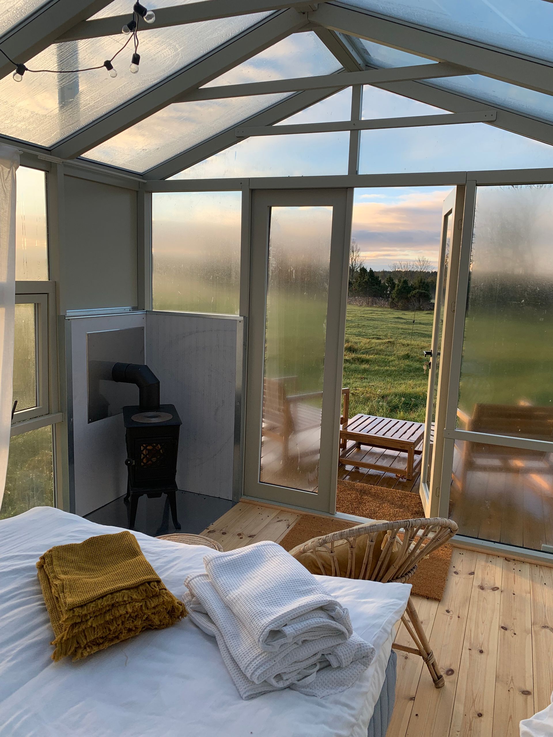 Cozy greenhouse bedroom with bed, chair, and wood-burning stove; open door to a grassy field.