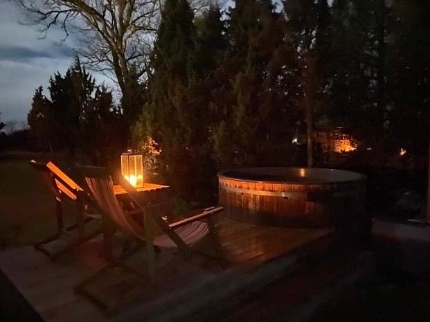 Wooden hot tub and two chairs lit by candlelight at dusk in a yard.