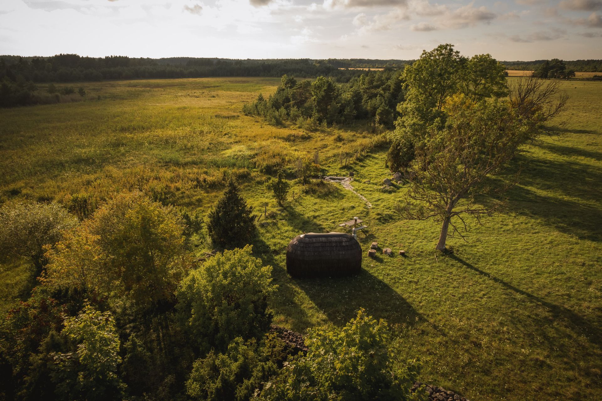 Golden-lit field with trees and a hay bale, a wooded area in the background under a cloudy sky.