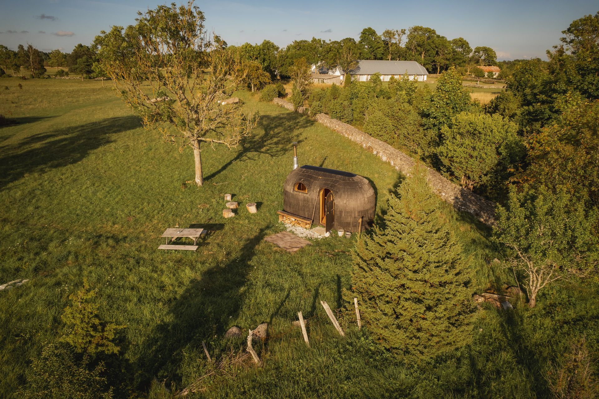 A grassy hillside with a small, rounded cabin. A picnic table and trees surround the structure.