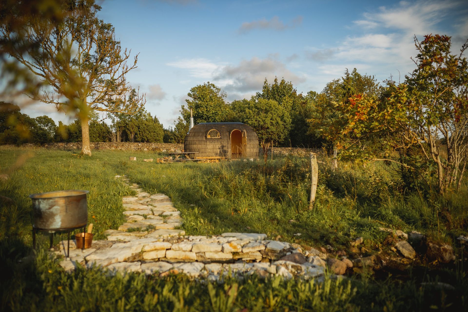 Stone hut in a field, with path, cooking pot, and surrounding trees under a blue sky.