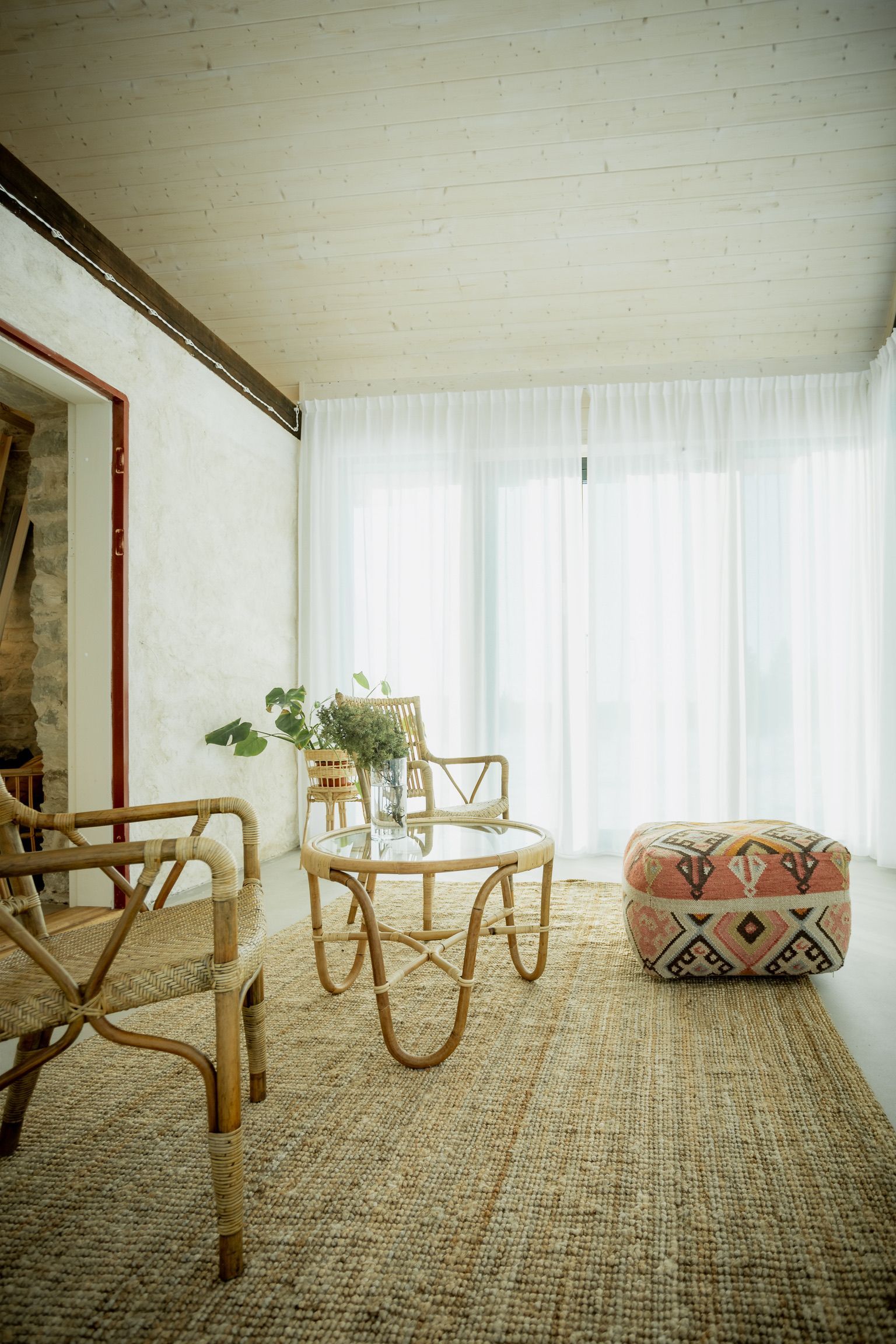 Living room with rattan furniture, a patterned ottoman, and a jute rug. Bright white curtains and walls.