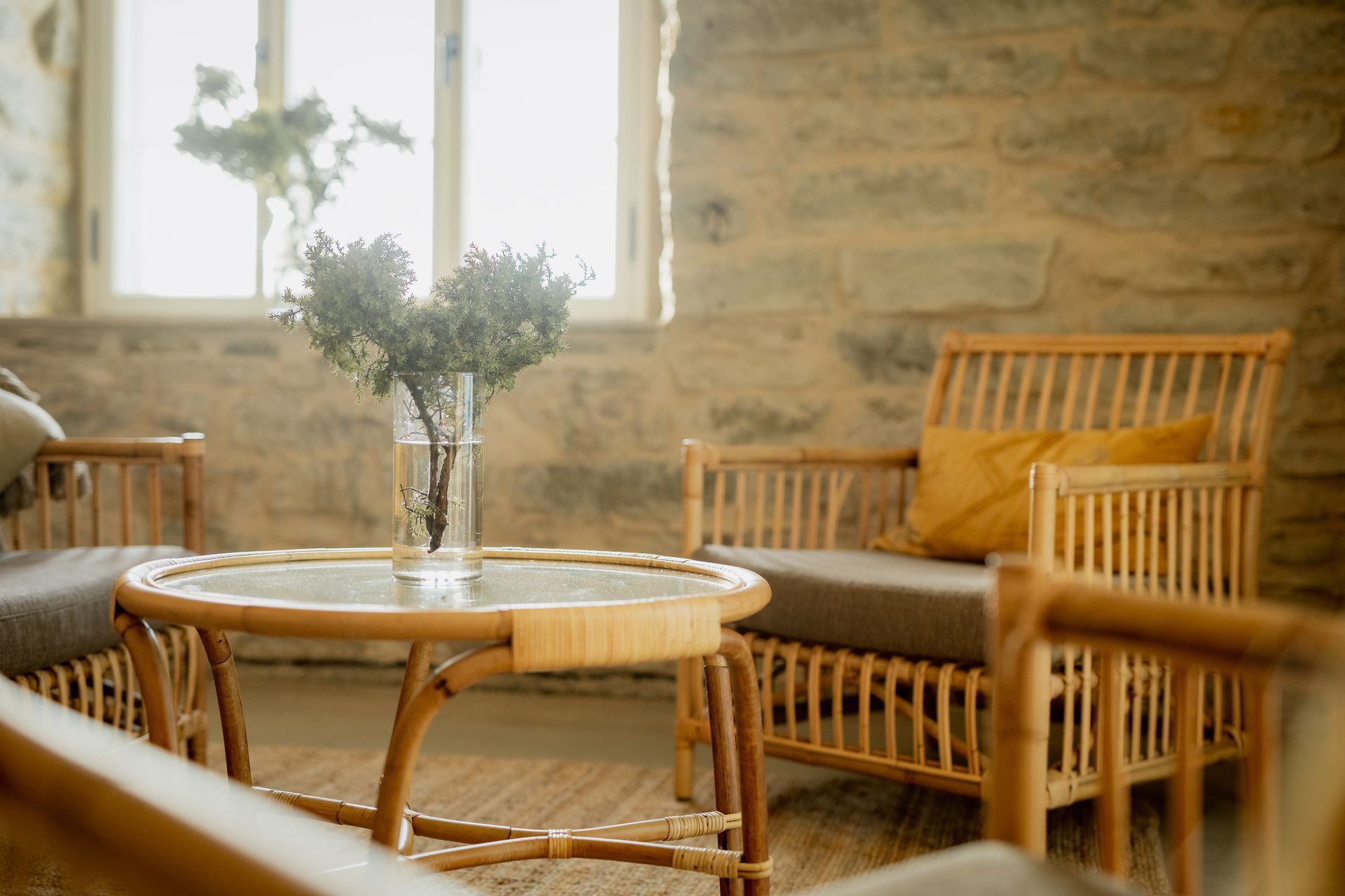 Rattan chairs and table in a room with stone walls, a vase of branches, and a window.