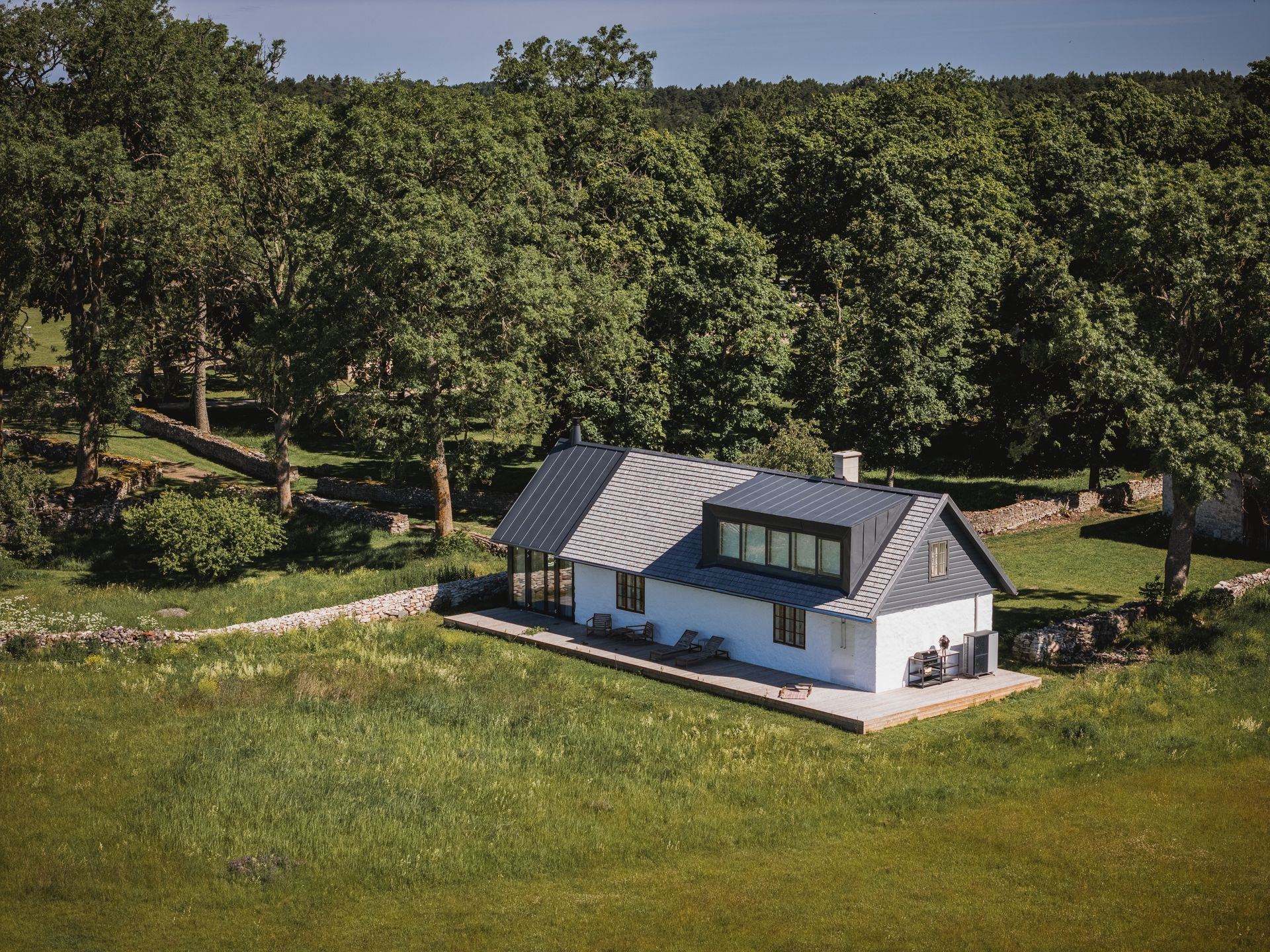 White house with dark gray roof, next to a stone wall, in a green field with trees.