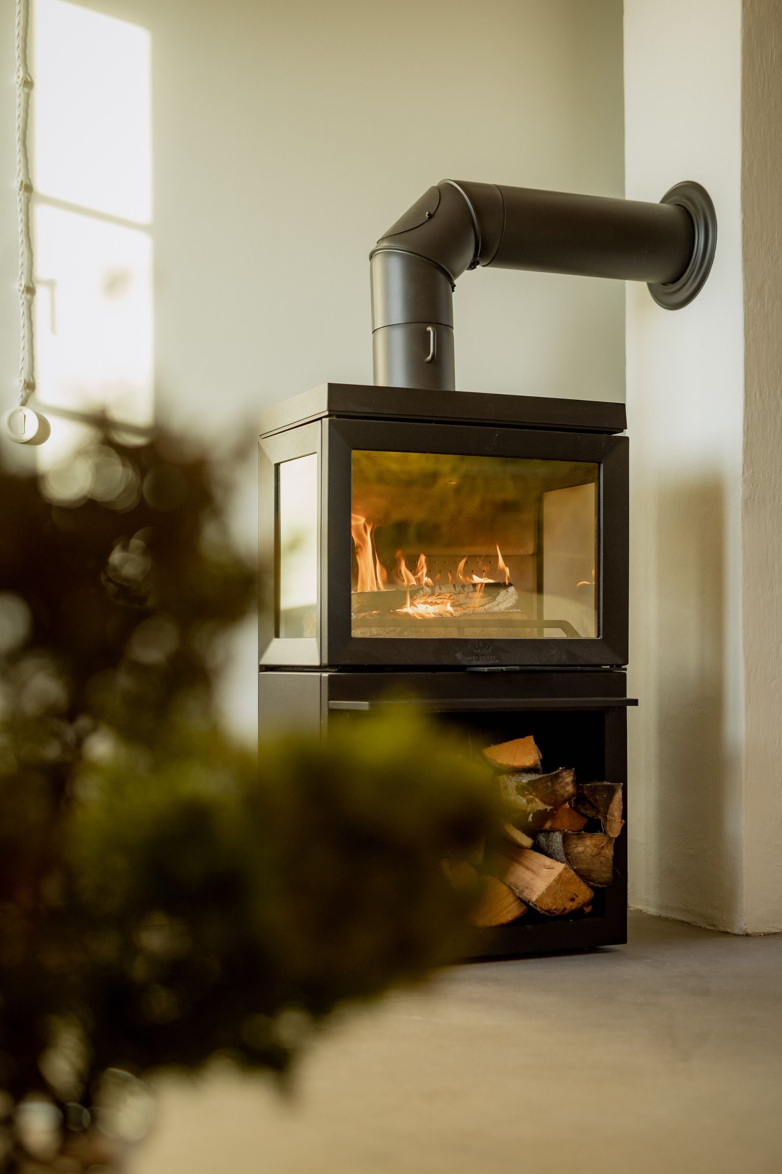 Modern fireplace with glowing flames and wood storage, black chimney pipe against a white wall.