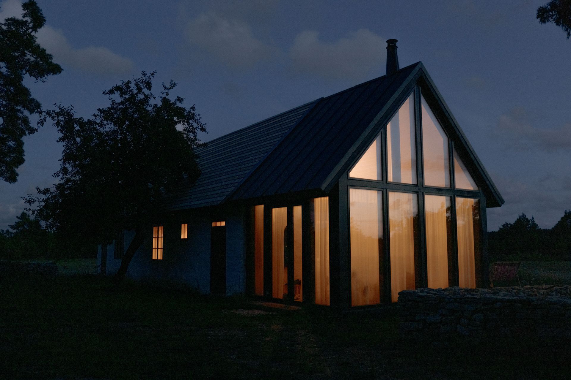 House with large windows lit from within at dusk; dark trees and sky surround.