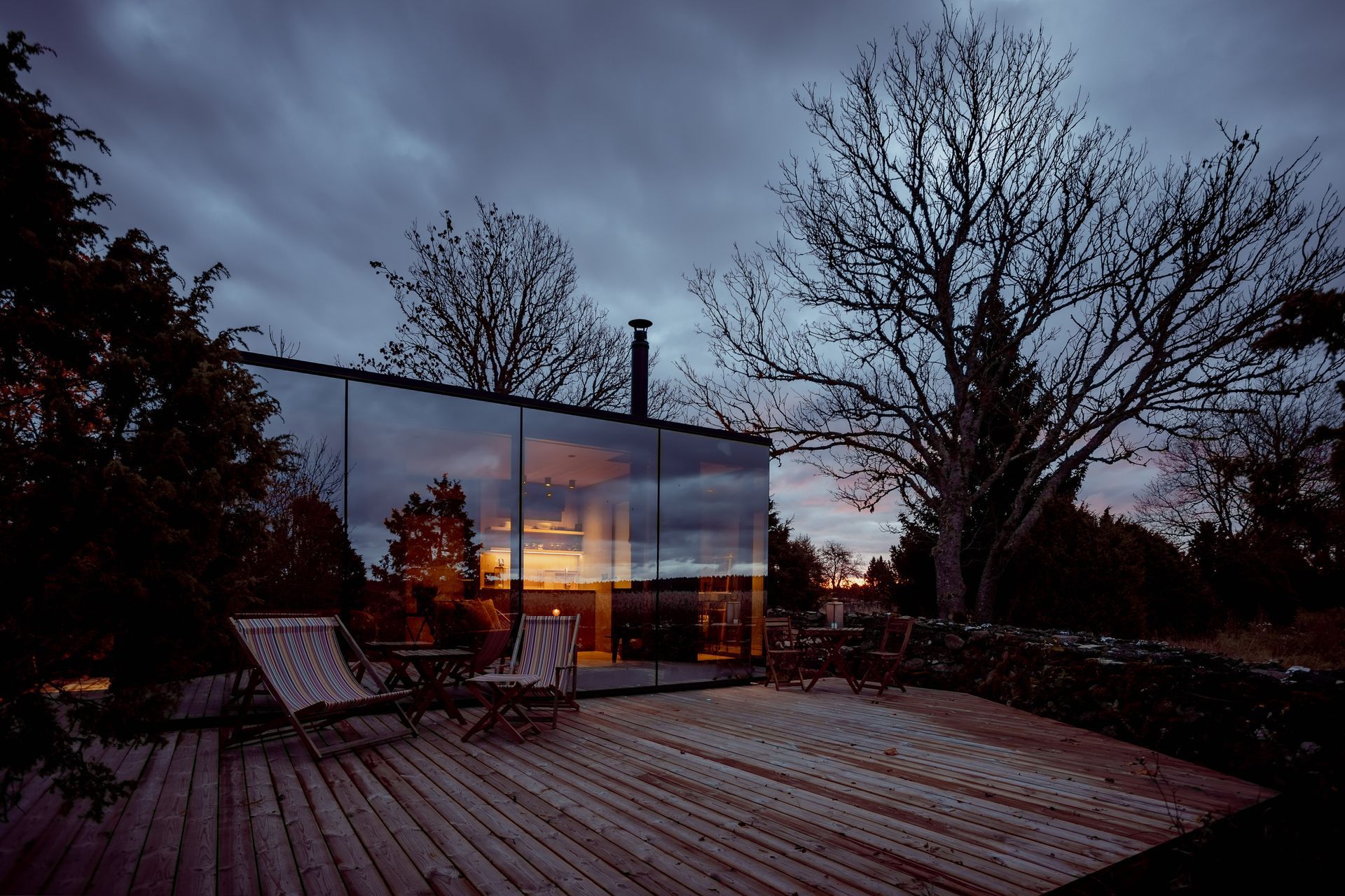 Glass-walled cabin at dusk, reflecting sunset. Wooden deck with two chairs, silhouetted tree, cloudy sky.