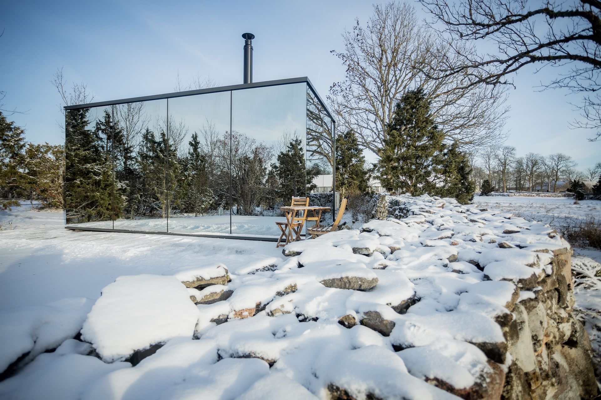 Mirrored cabin in snowy field reflects trees and sky. Stone wall in foreground.