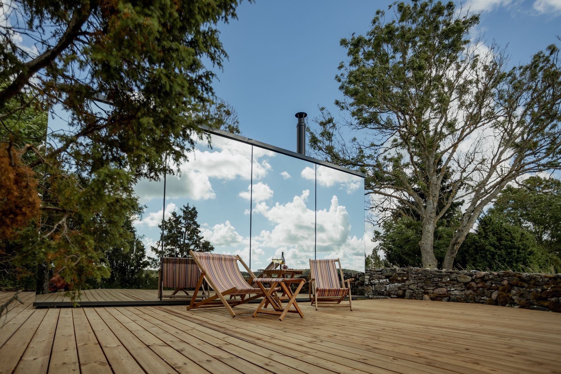 A modern cabin with mirrored walls reflects the sky, with two chairs on a wooden deck.
