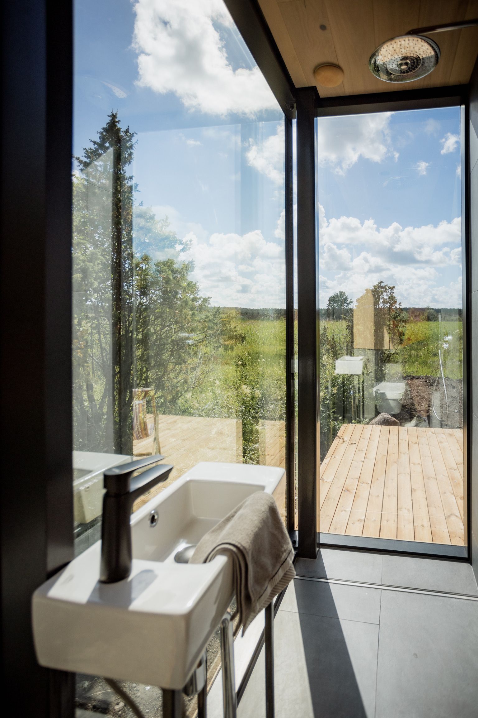 Bathroom with a white sink, black faucet, and a view through a large window of greenery and a blue sky with clouds.