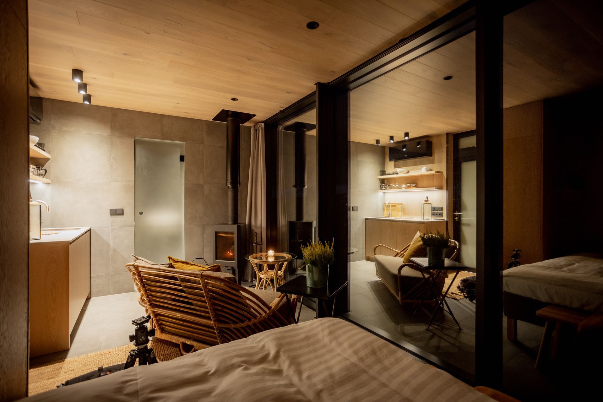Cozy cabin interior with bed, wicker chair, small fireplace, and a kitchenette visible through glass. Warm lighting.