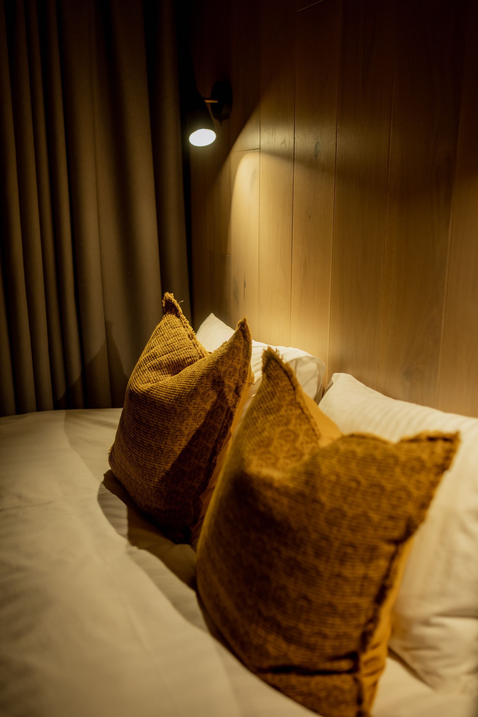 Bed with yellow decorative pillows, under a small reading light against a wood-paneled wall.