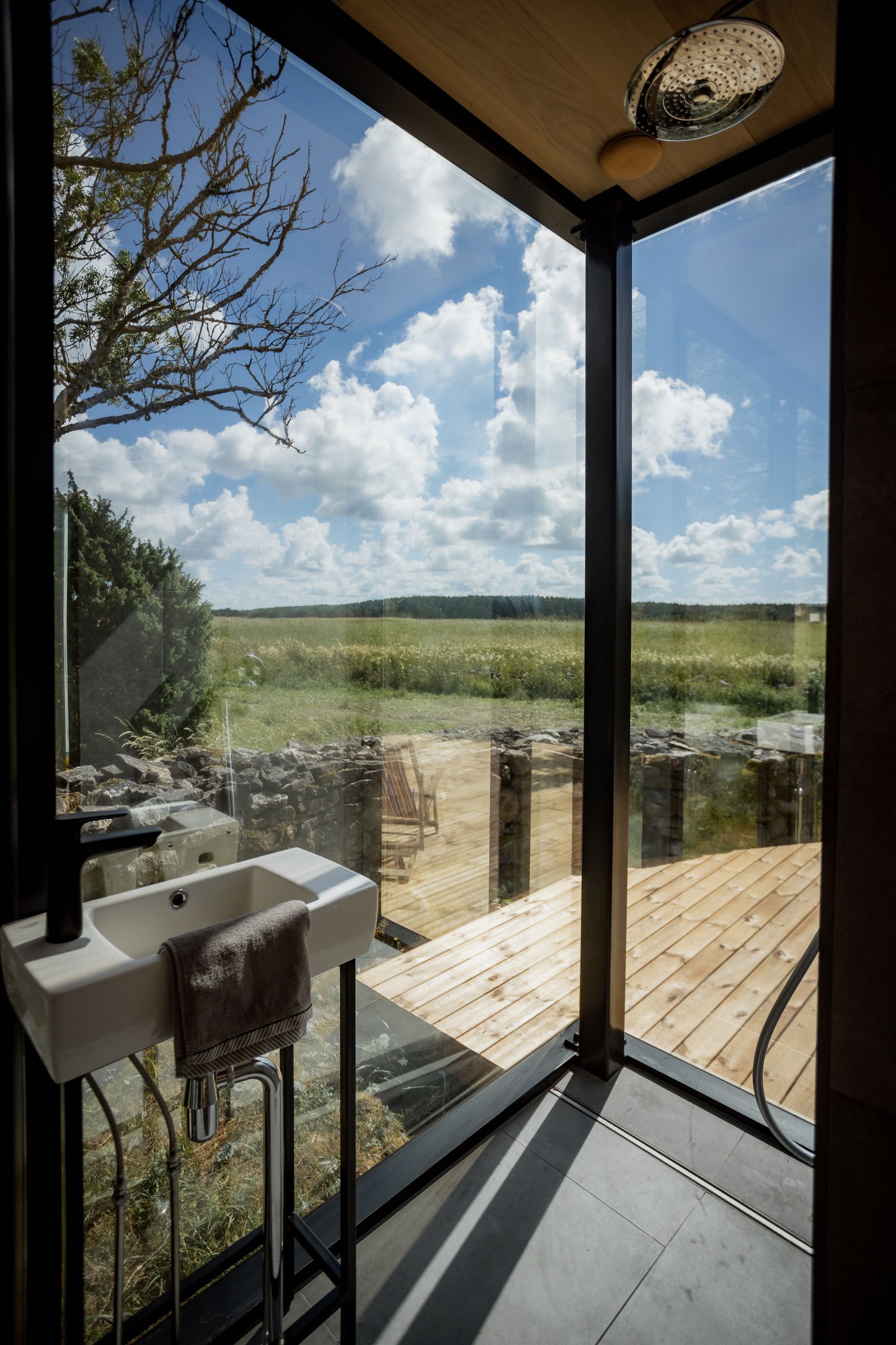Bathroom with large window overlooking a field on a sunny day. Sink, showerhead visible.