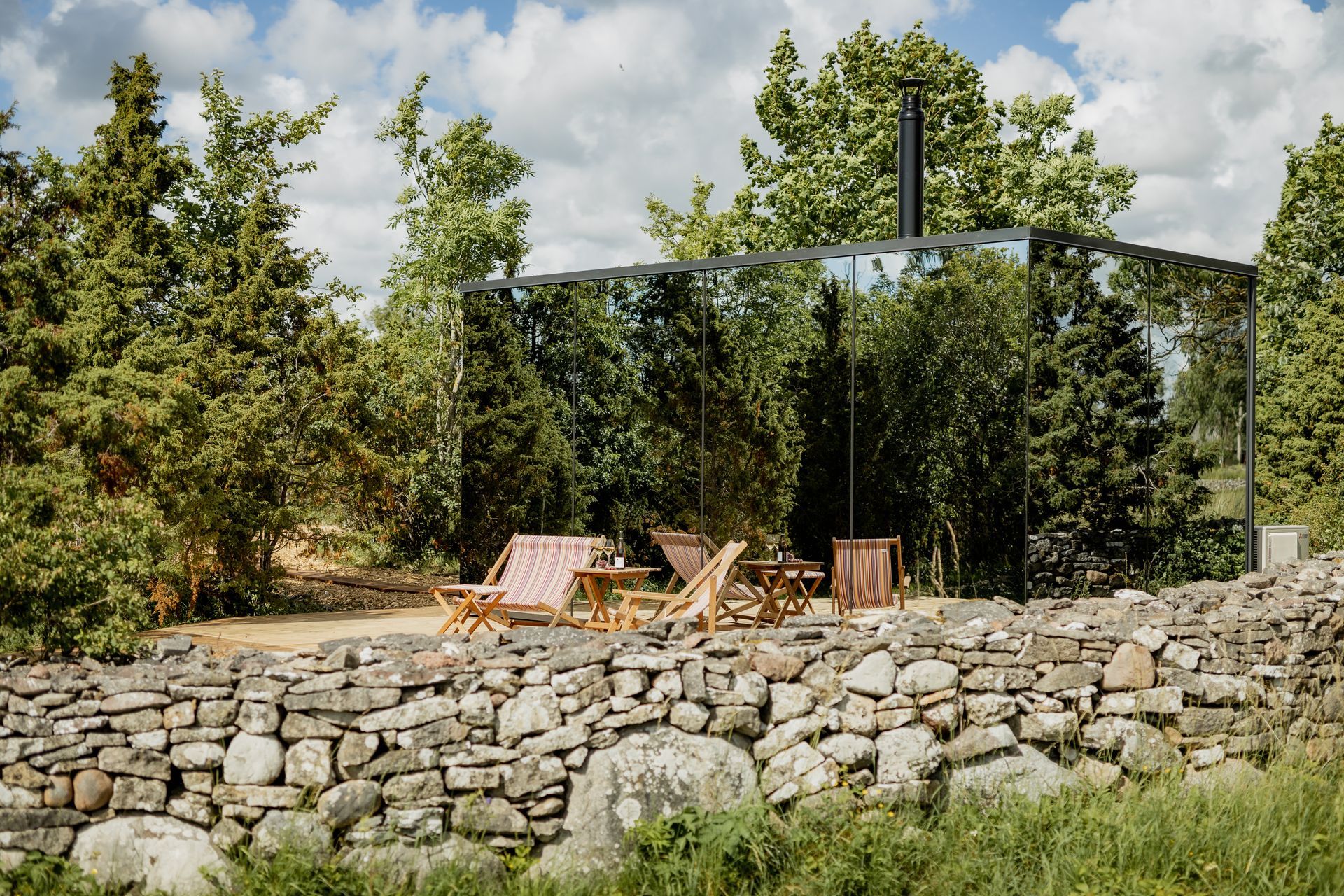 Mirrored cabin reflecting surrounding trees, with stone wall and deck chairs.