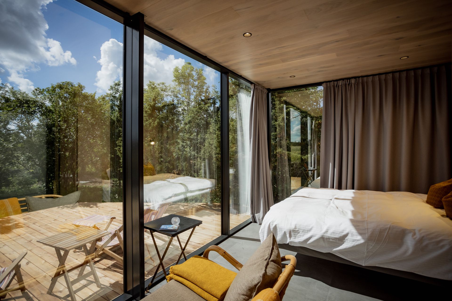 Bedroom with large glass windows overlooking a wooden deck and trees. Bed is white with yellow accents.