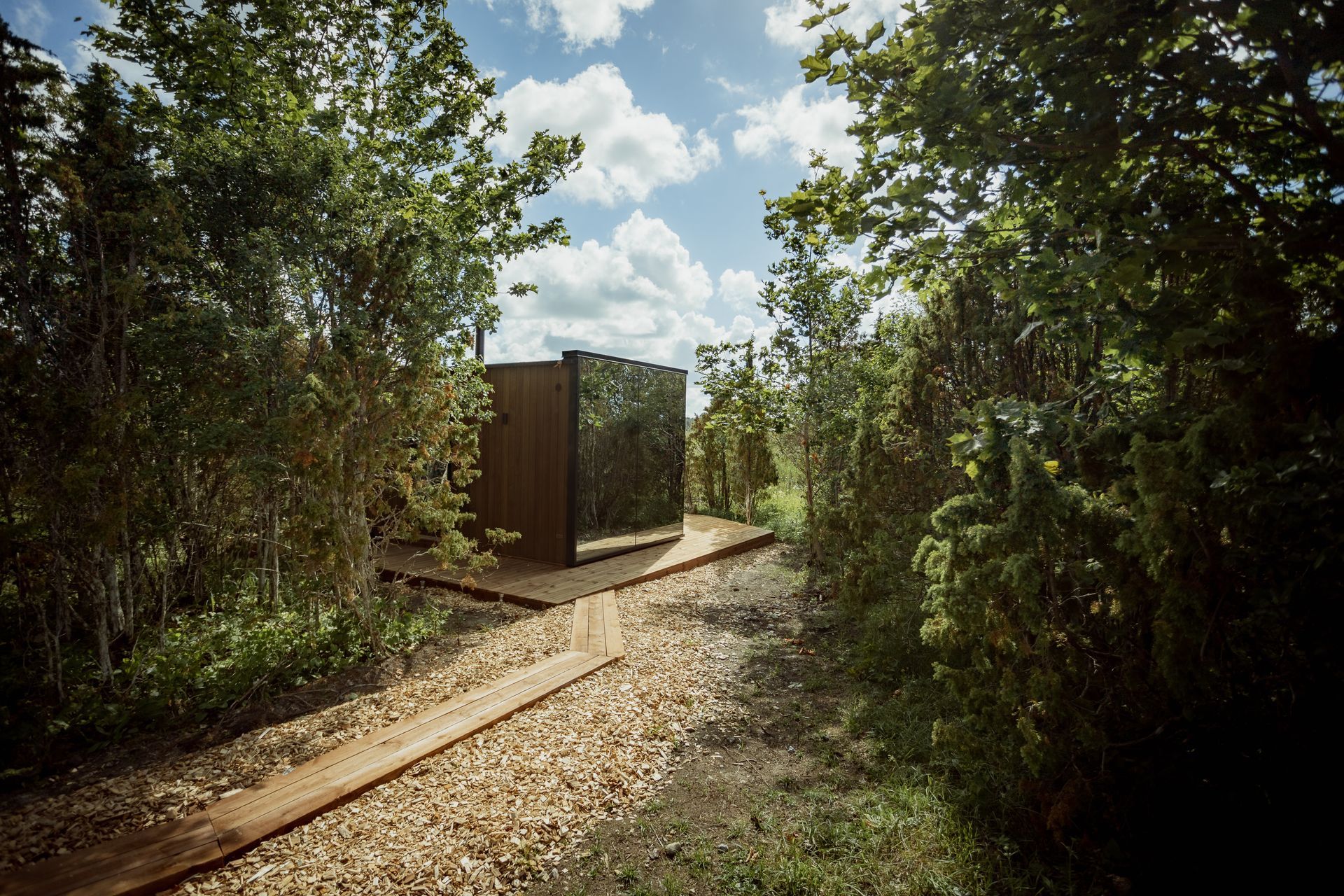 Wooden path leads to a modern, mirrored structure in a lush, green forest under a cloudy sky.