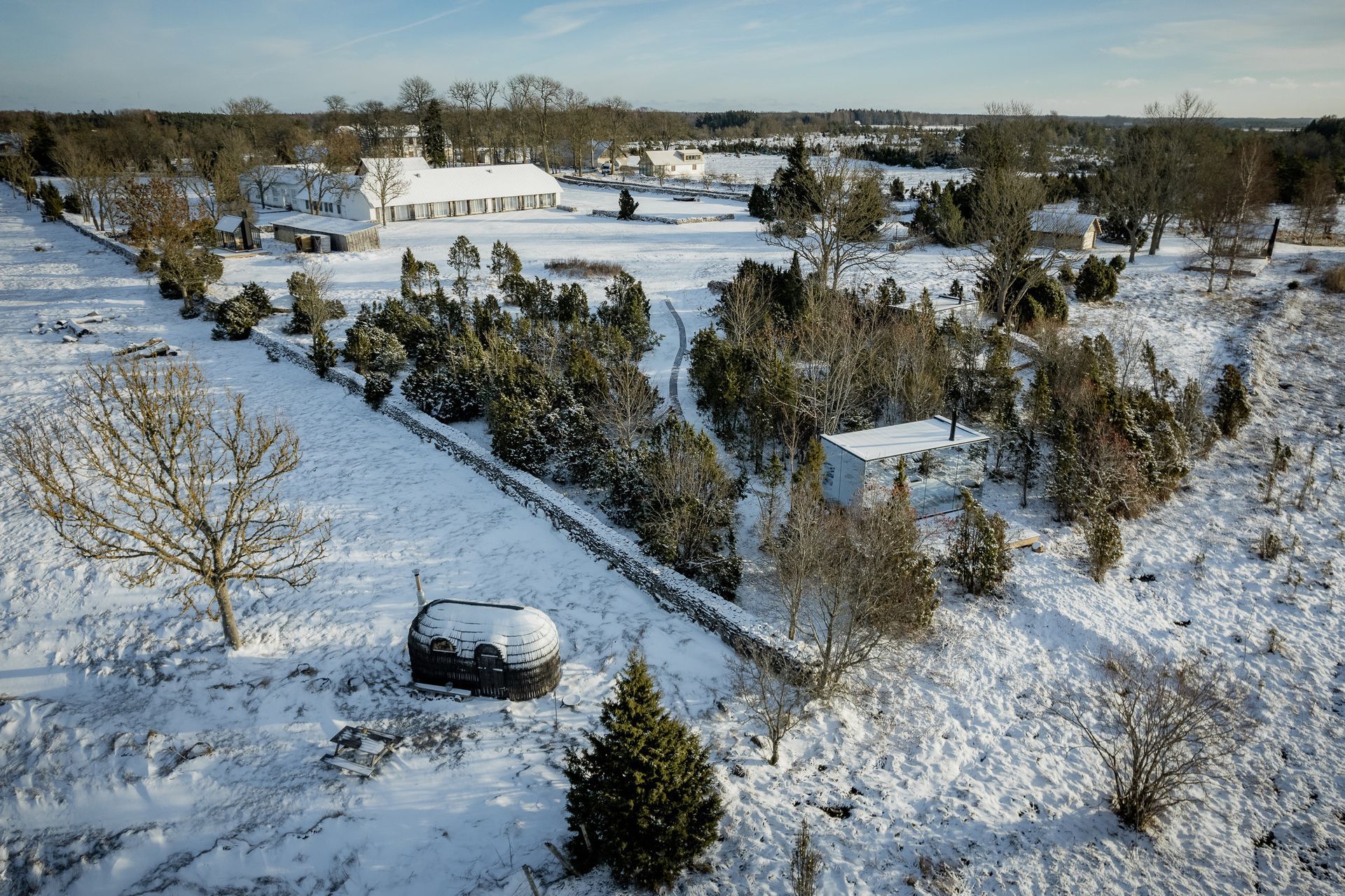 Snowy rural landscape with buildings, trees, and a stone wall.