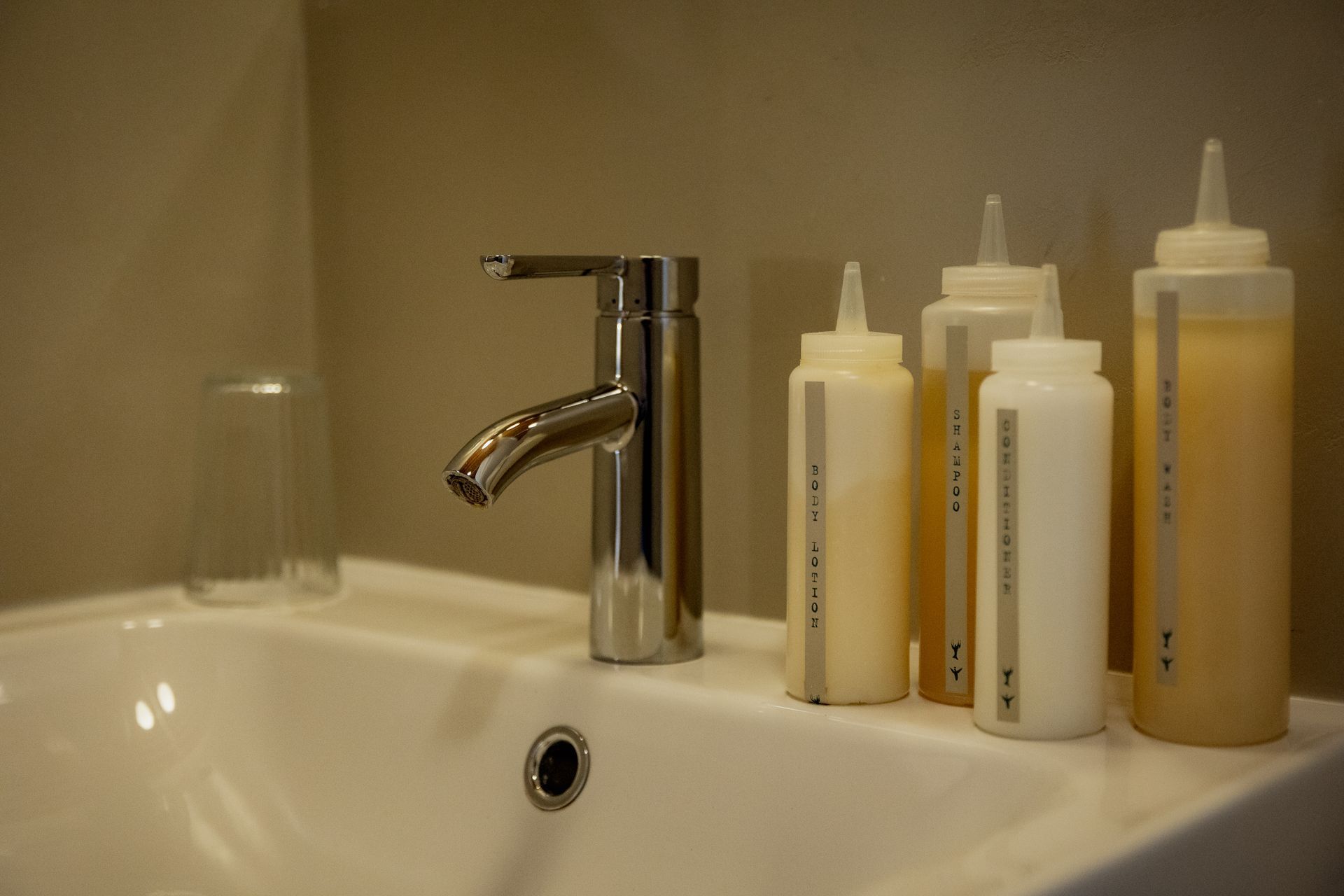 A bathroom sink with a chrome faucet, clear glass, and squeeze bottles containing liquid.