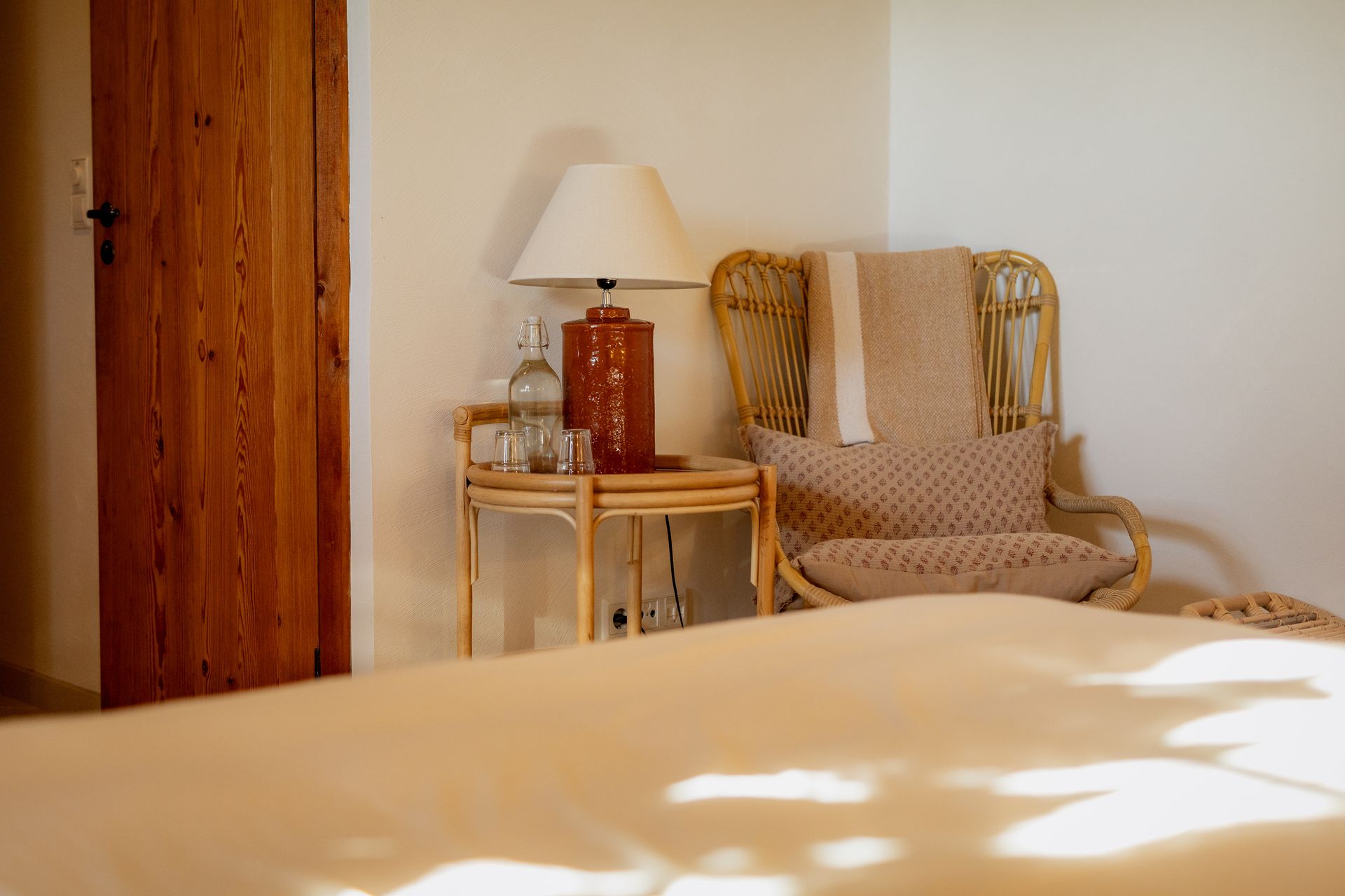 Cozy bedroom corner with rattan chair, side table, lamp, and wooden door. Neutral tones, soft lighting.