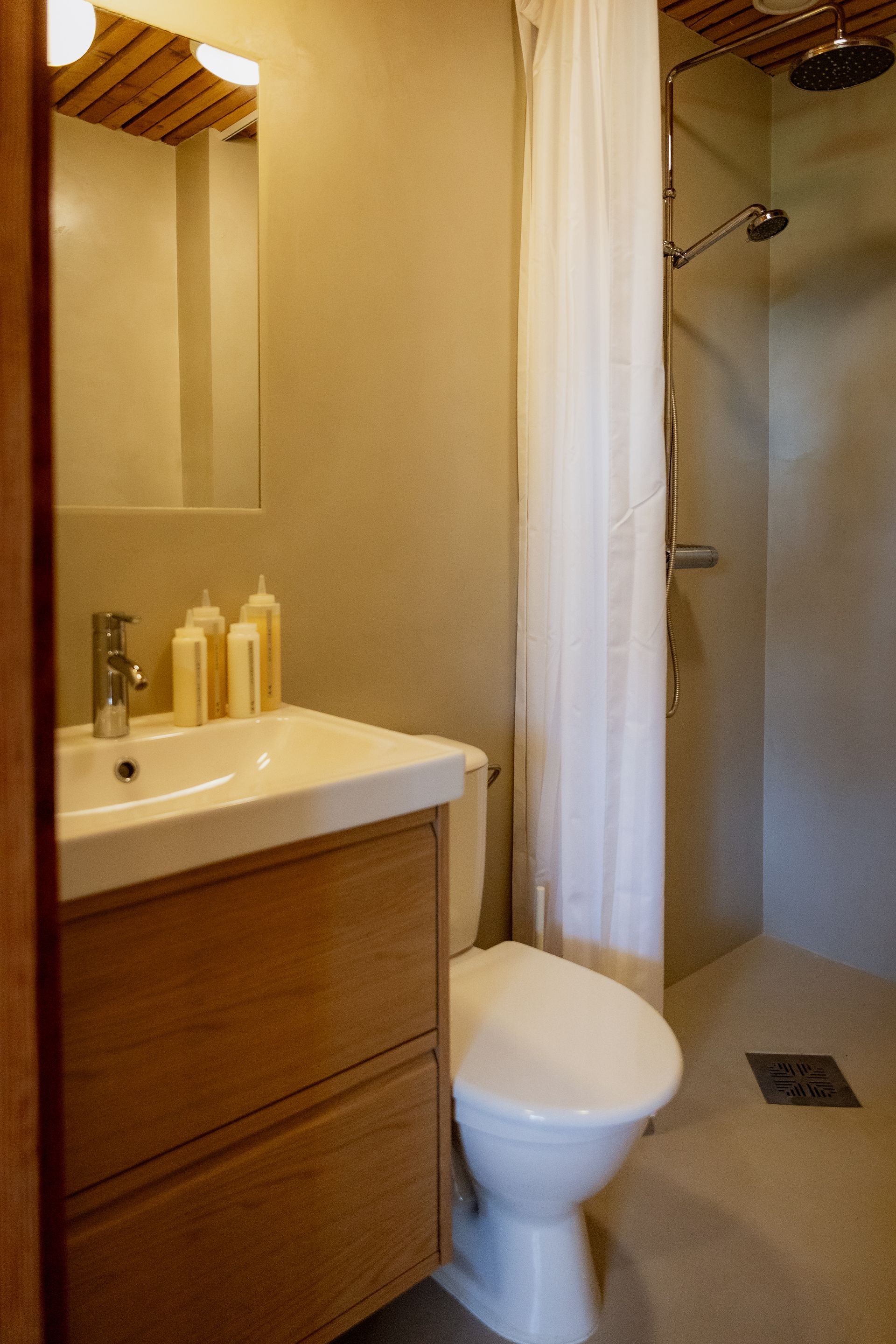 Bathroom with wooden vanity, toilet, and shower stall. Beige walls, white shower curtain, and natural light.