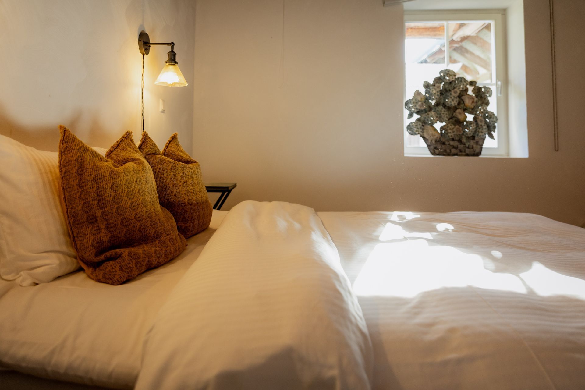 Cozy bedroom with white bedding, two brown pillows, a lamp, and a window with light.