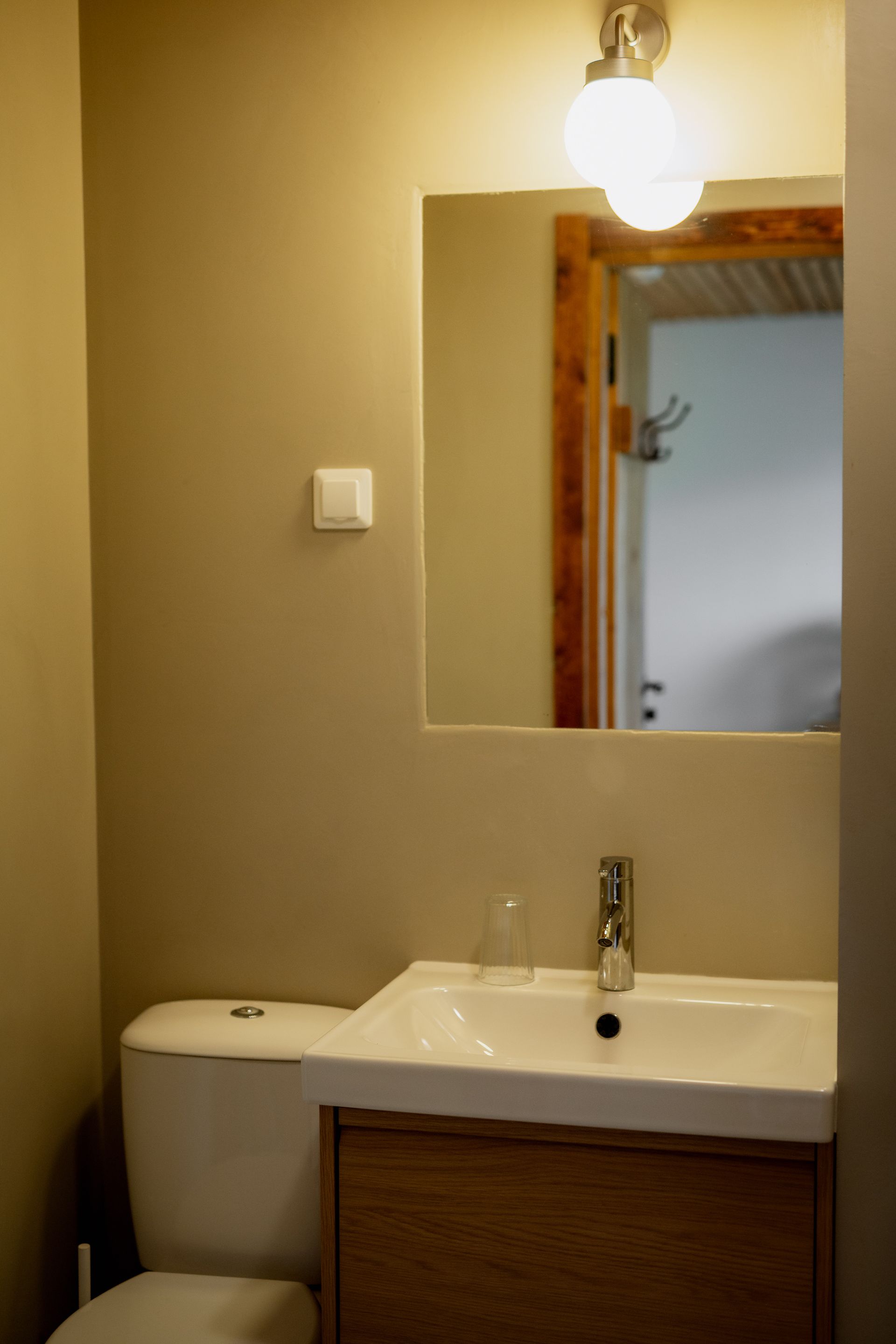 Bathroom with toilet, sink, mirror, and light fixture. Neutral beige walls and wooden accents.