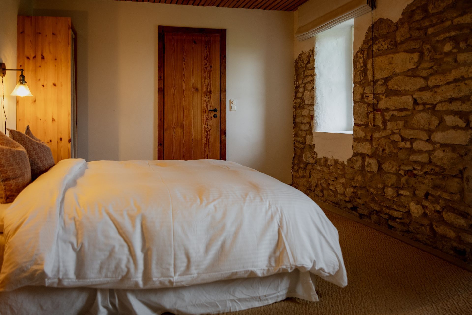 Bedroom with a bed, wooden door, exposed stone wall, and window with sheer curtain.