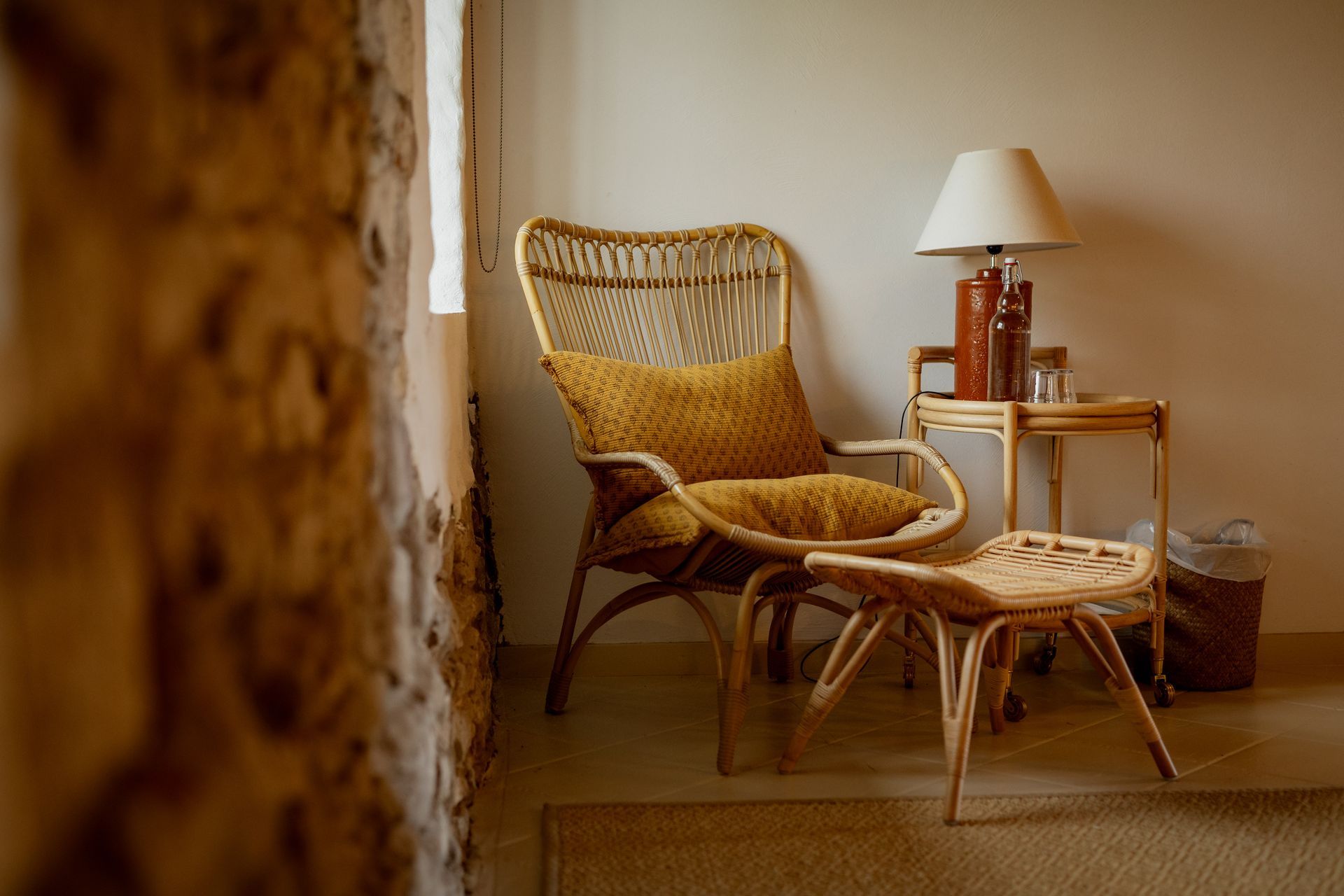 Wicker chair with cushion, lamp, stool, and small table by a stone wall.