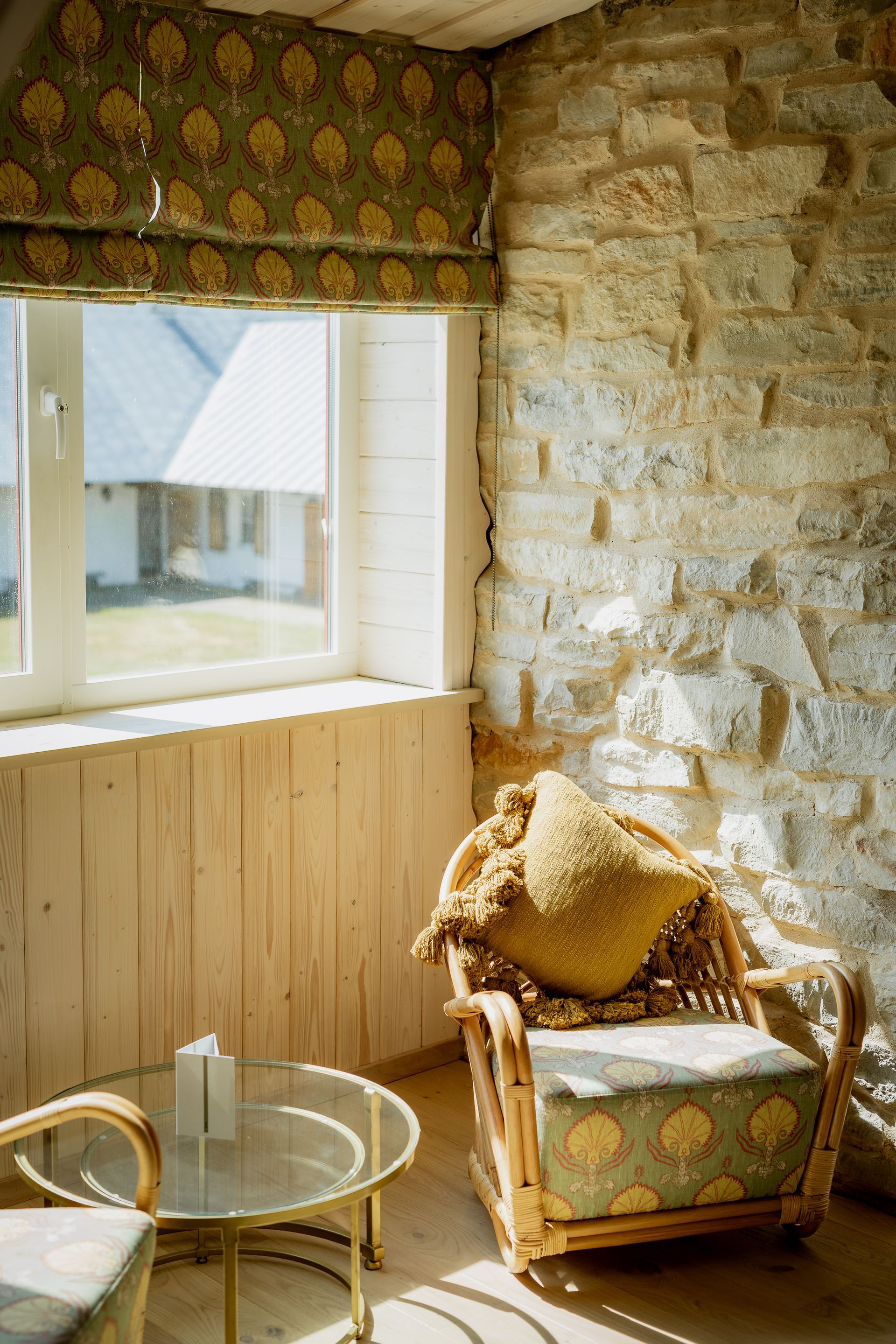 Cozy sunroom with wicker chair, patterned cushions, small table, and stone wall; natural light from window.