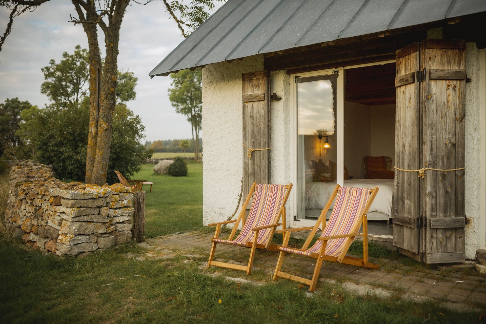 Two folding chairs sit outside a small white building with wooden shutters and an open door. Green field in the background.